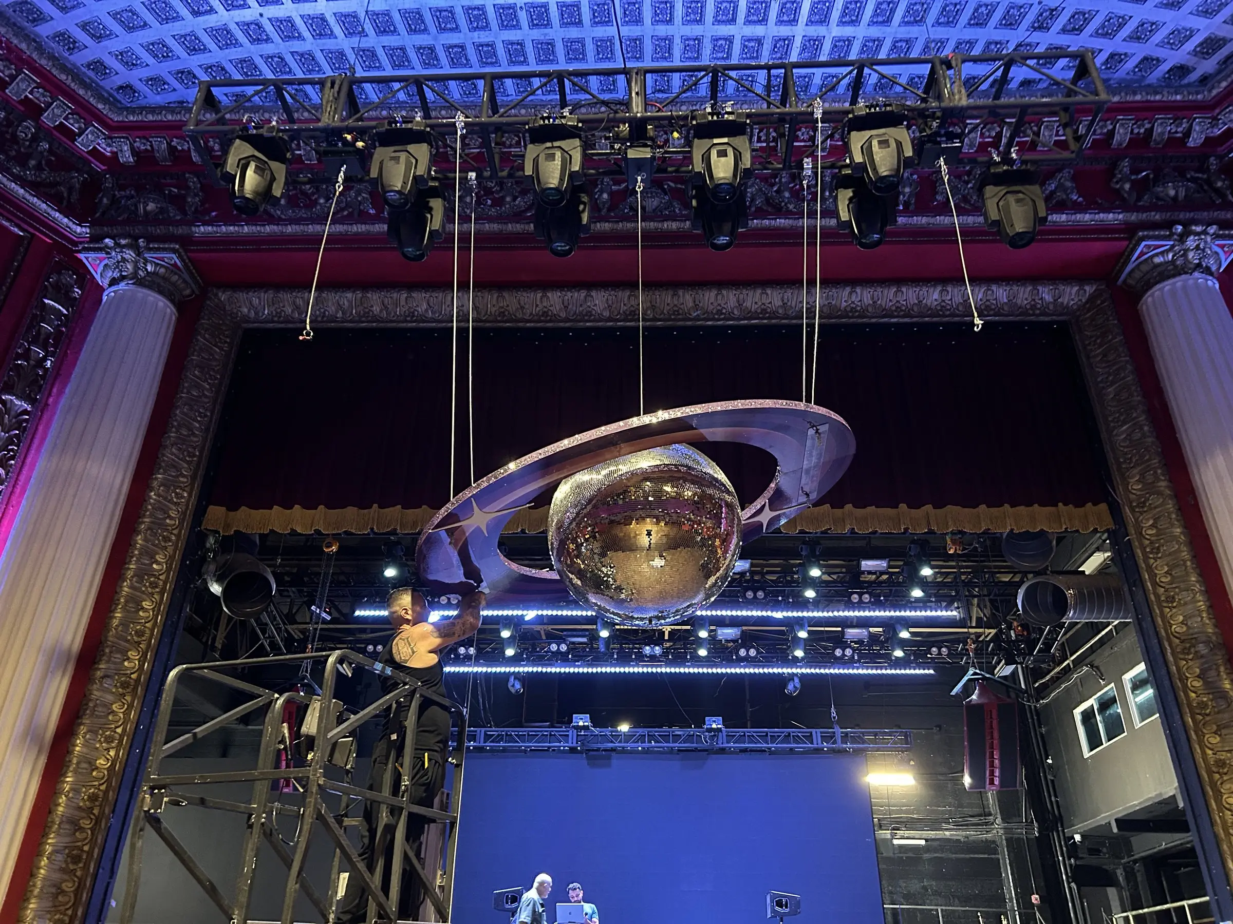 Looking up at a mirror ball inside a wide ring, hung from the lighting bars under the ornate ceiling.