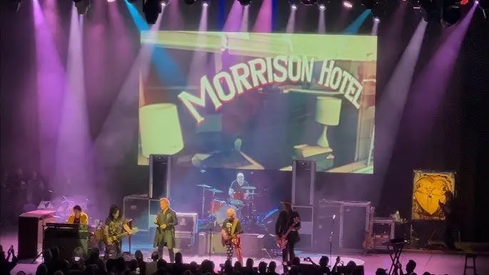 The band playing under purple and blue beams, a wide screen behind them showing a Morrison Hotel sign, and the crowd in silhouette across the foreground.
