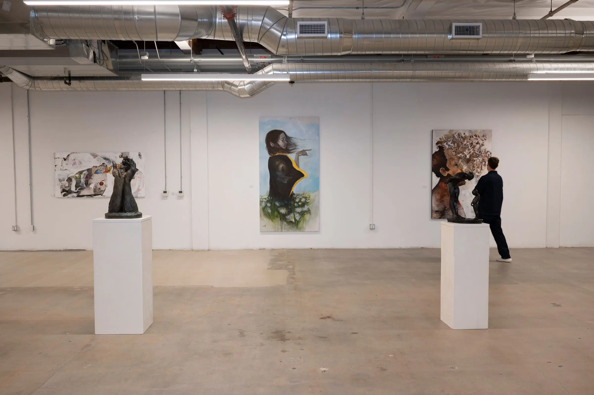Two bronze figures on white plinths in front of three paintings on a gallery wall, with one guest walking out of the frame at the right.