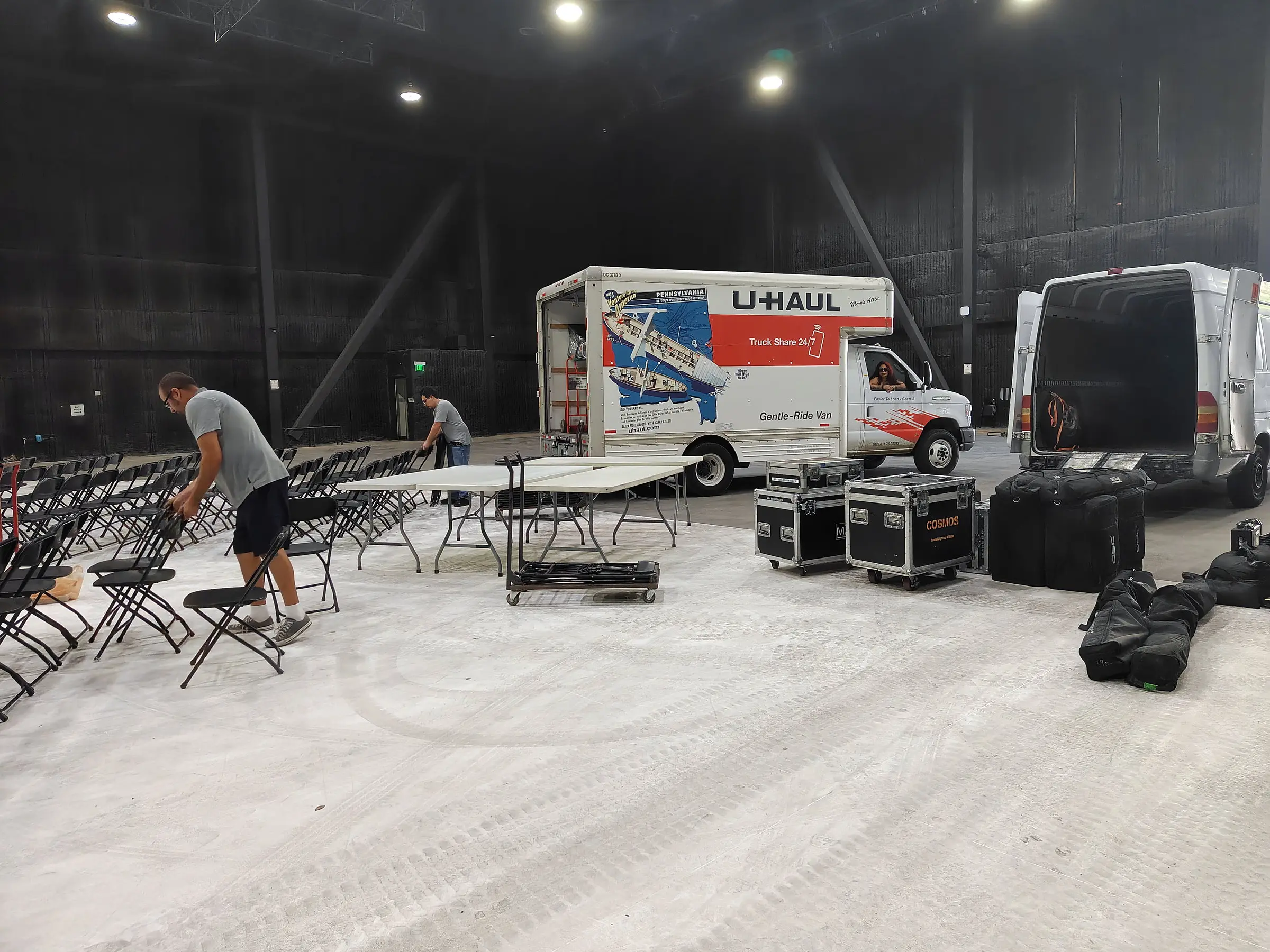 The soundstage during load-in, a rental box truck and a cargo van parked on the concrete, road cases along the wall, and crew setting rows of folding chairs.