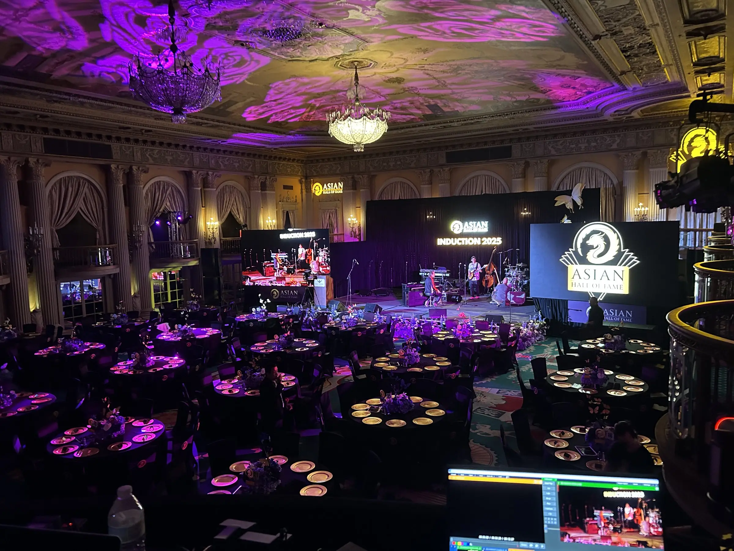 The same ballroom from the balcony, chandeliers overhead and a band playing on the stage beside a logo screen.
