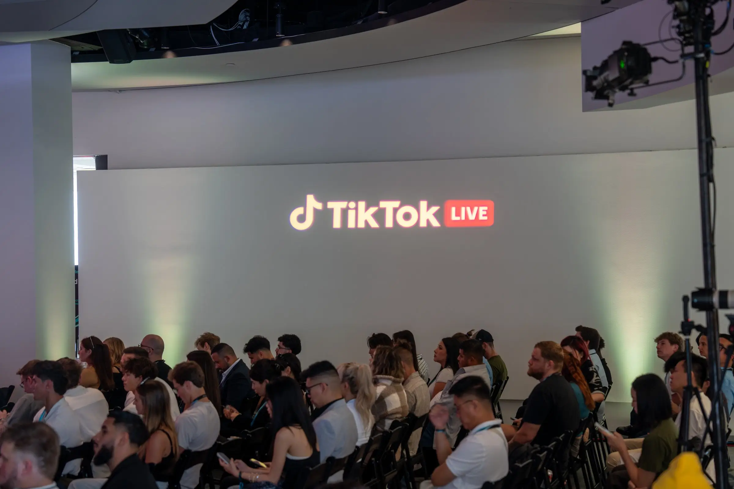 The seated audience along a white wall with the TikTok Live logo glowing above them and uplights washing the wall green.