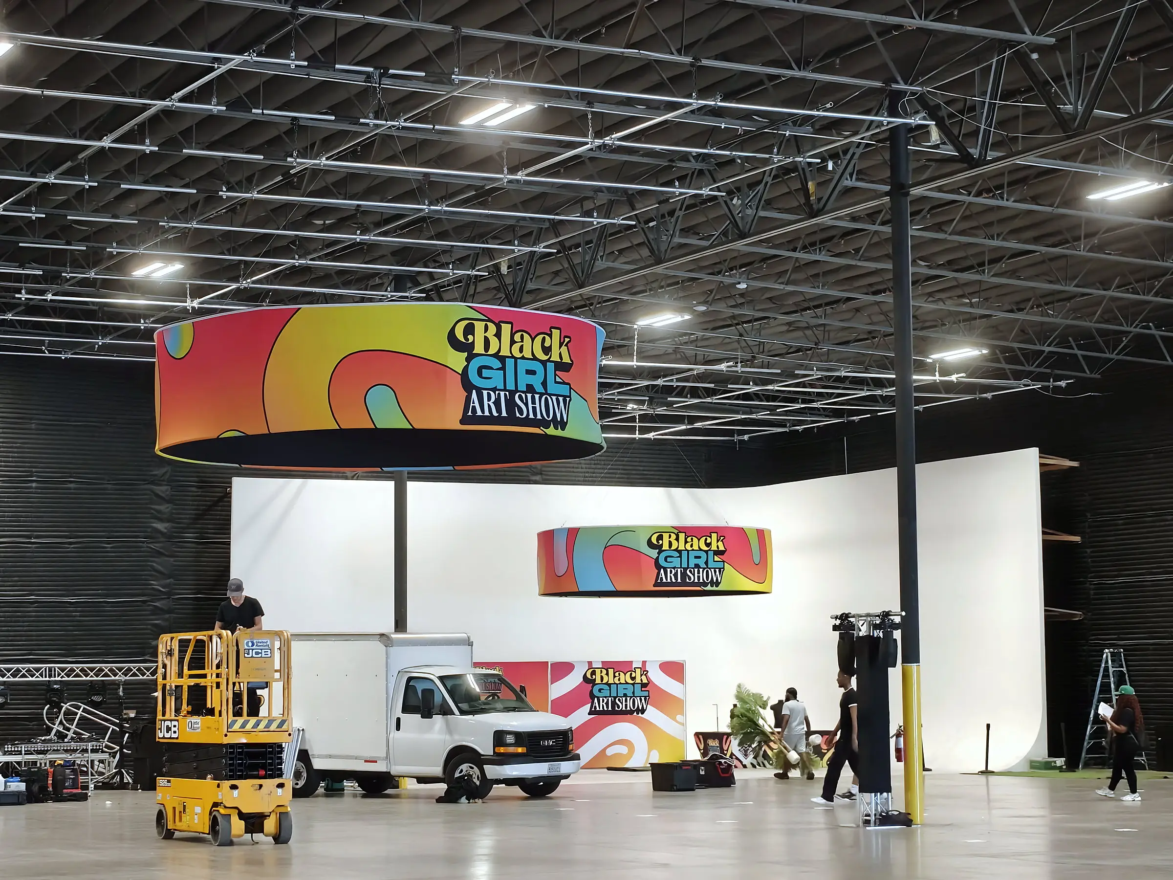 The build with a scissor lift and a box truck on the floor, both round Black Girl Art Show banners hung, and crew dressing the white wall behind them.