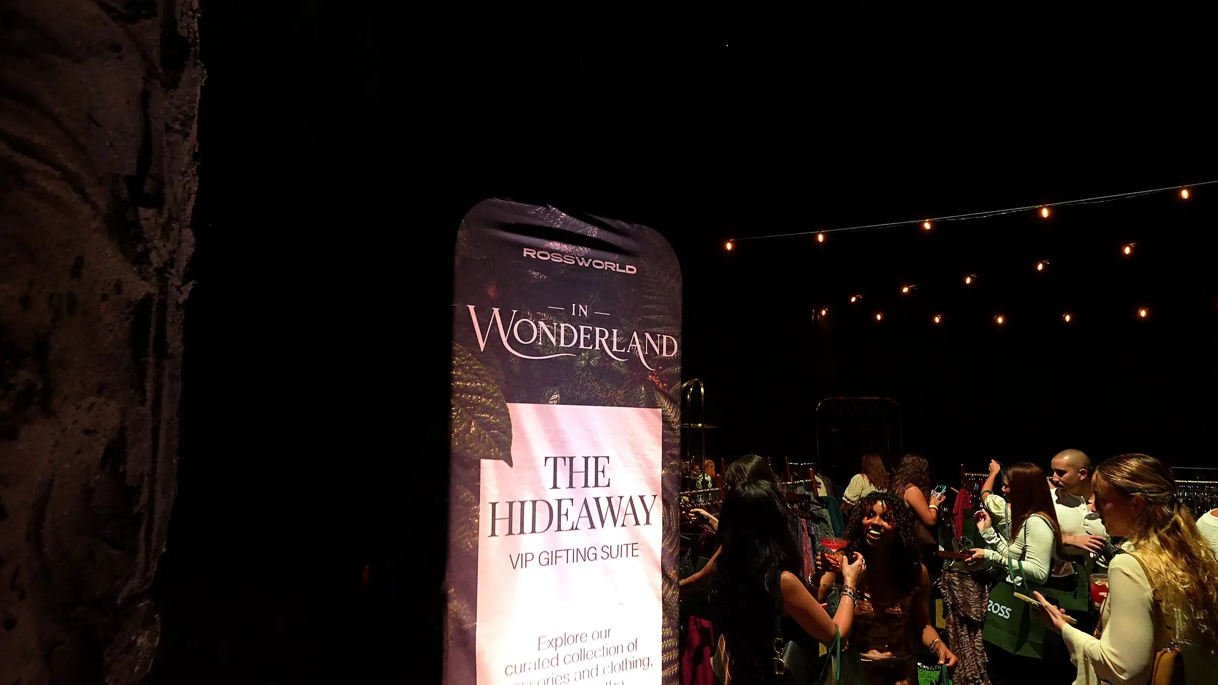 The Rossworld In Wonderland banner for The Hideaway VIP gifting suite, lit against the dark beside the rock wall, with guests at the racks under string lights beyond.