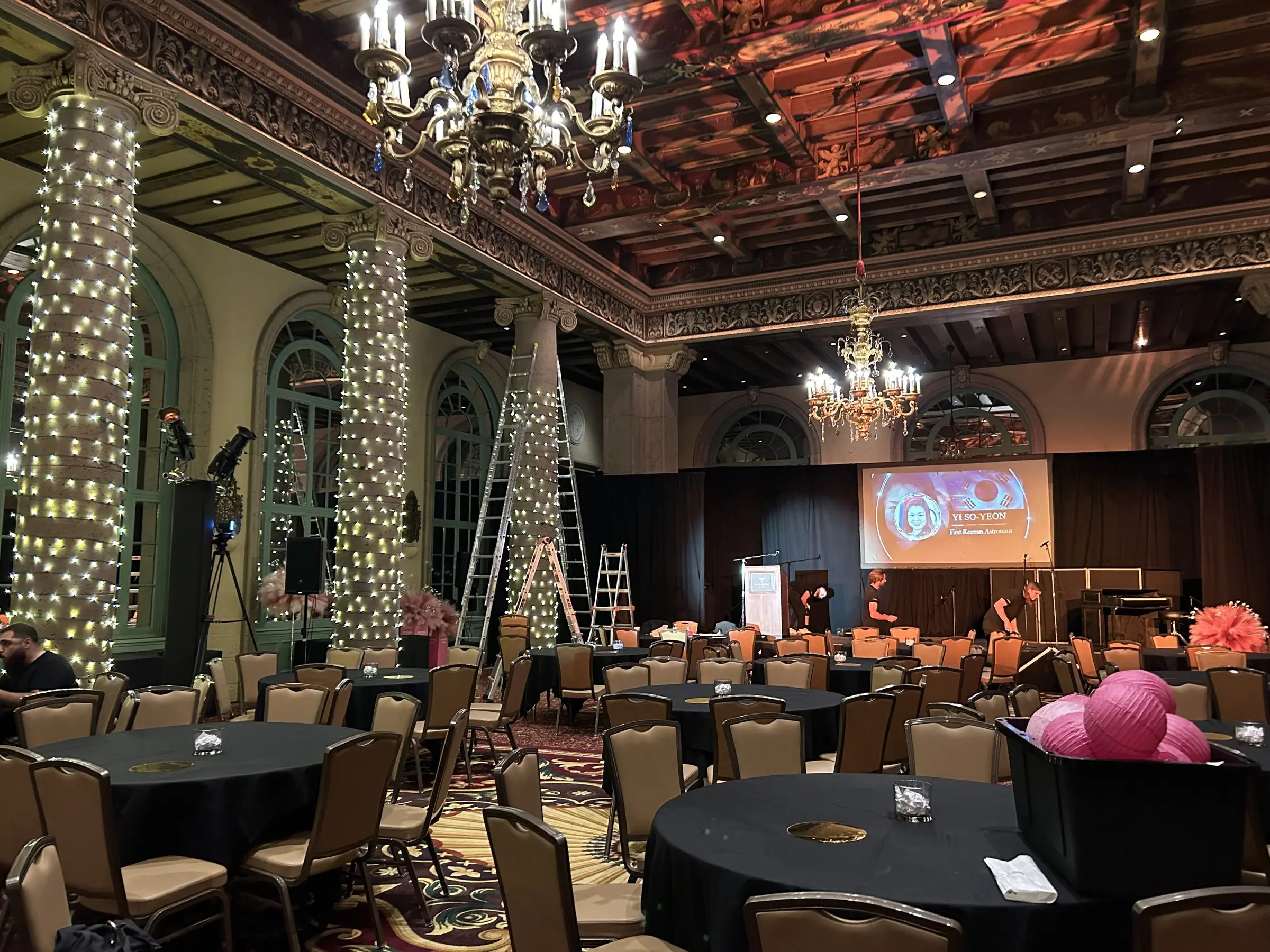 The same ballroom during setup, ladders against the columns while crews dress them in lights and the stage screen glows at the far end.
