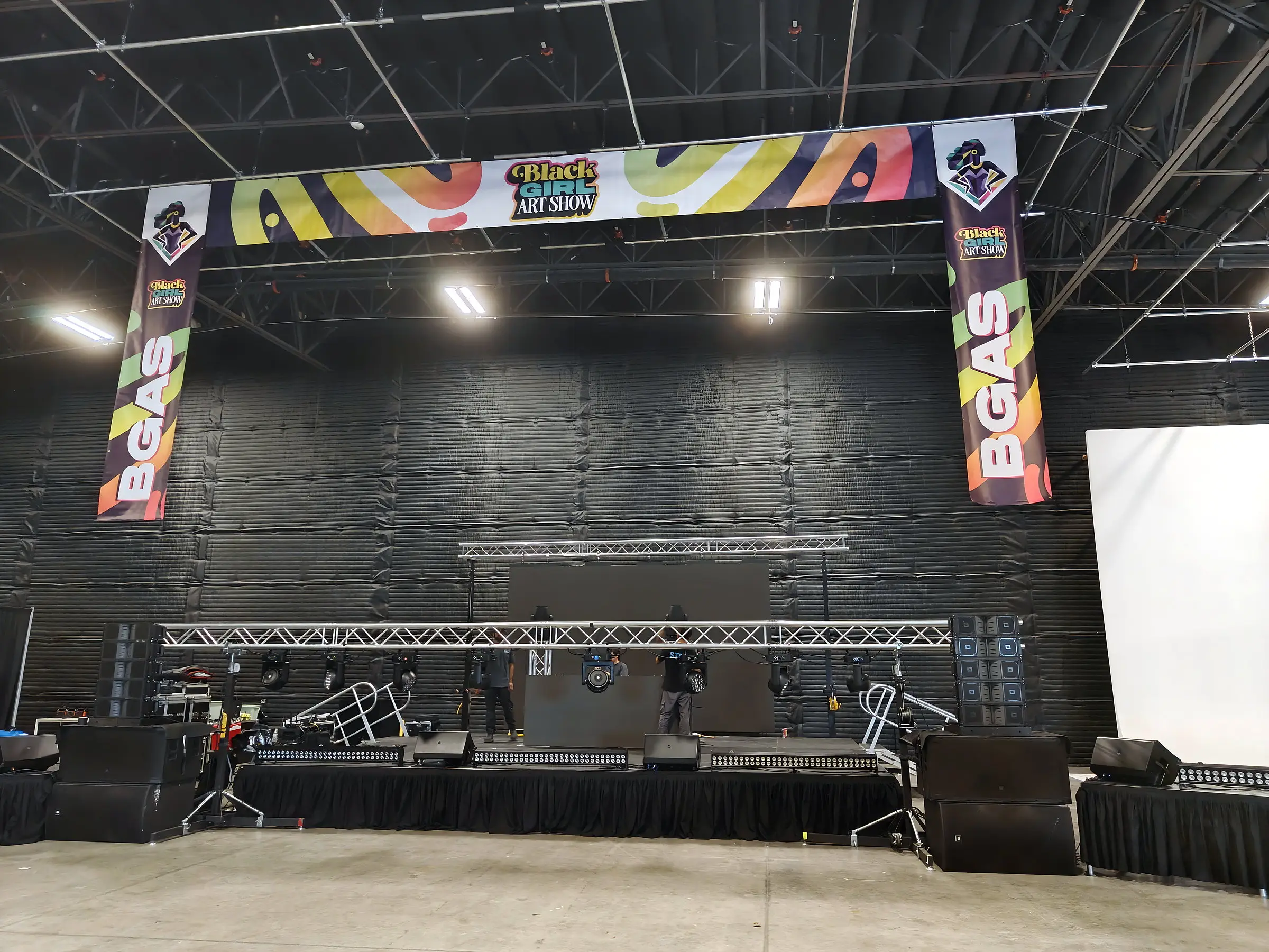 The stage during build, a truss of moving lights over the deck, line arrays at both sides, and a printed header banner with BGAS drops hung above it.