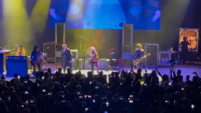 The same stage lit warm, a singer at the microphone in front of the band, amp stacks at either side, and the crowd holding up phones along the front.