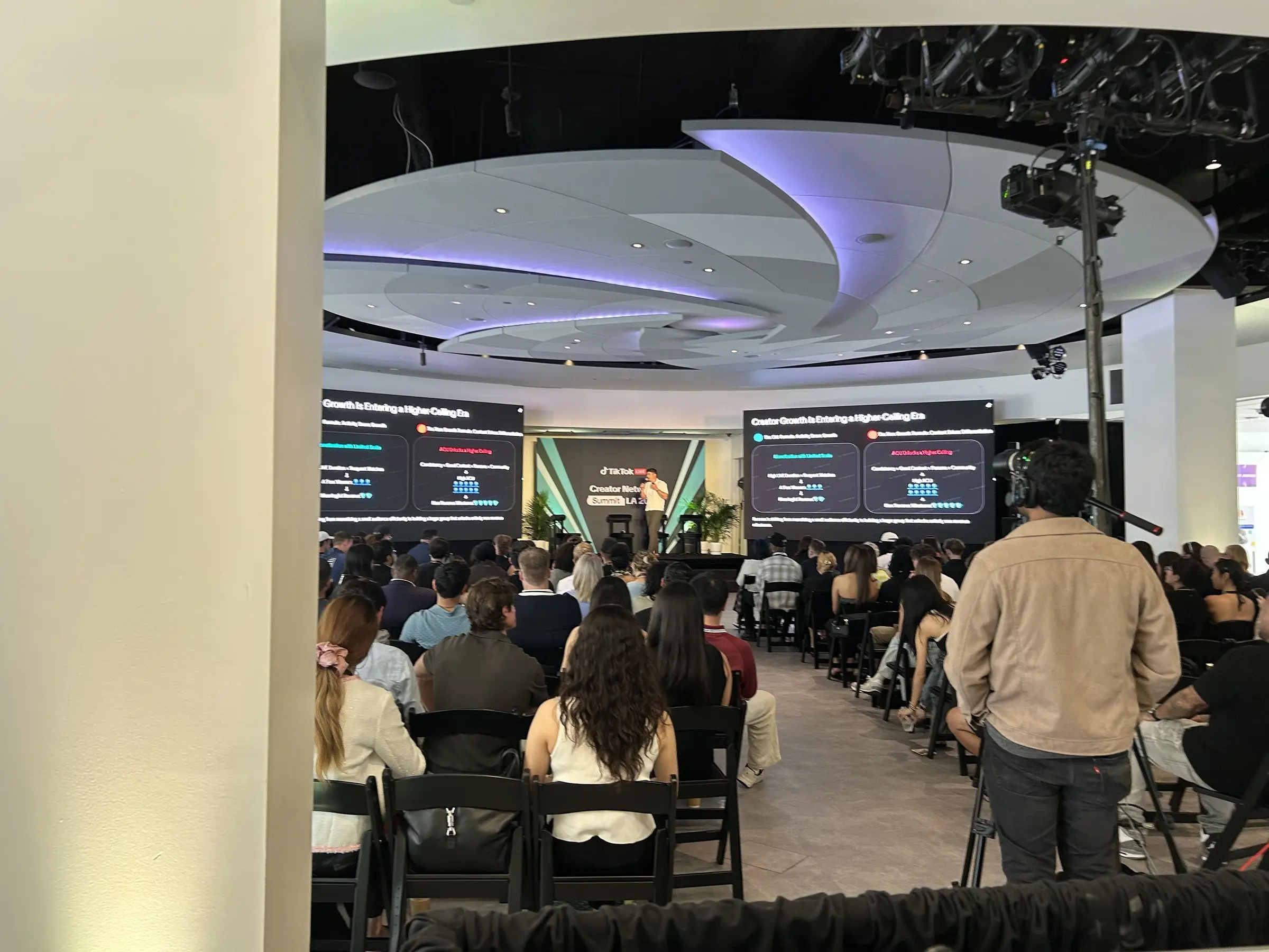 The room in session, seated guests filling the floor under the curved ceiling with the screens lit at the front.