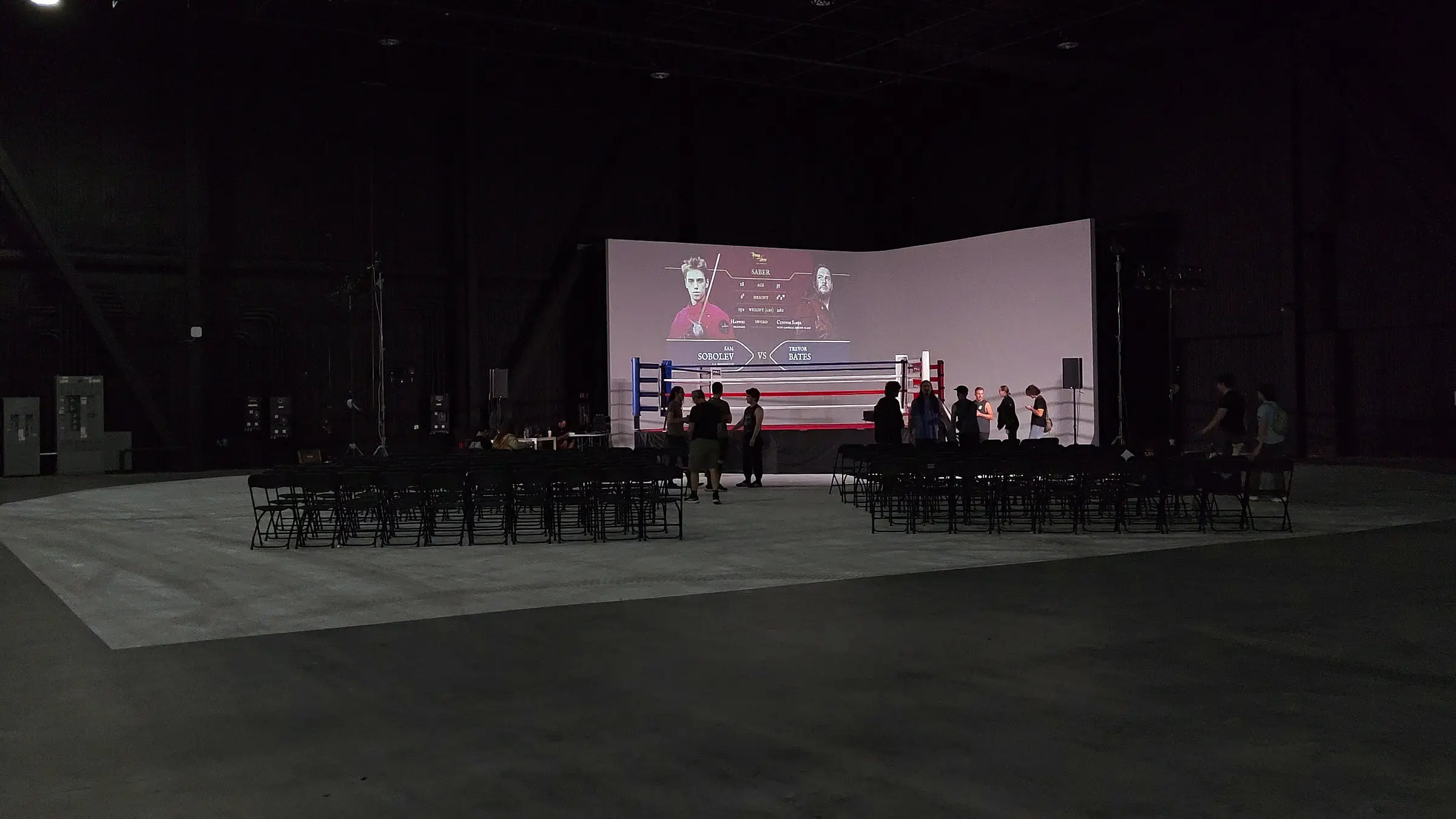 The built room from the back, the ring under a bout card projected across the white cyclorama, with empty chair rows on the concrete floor.