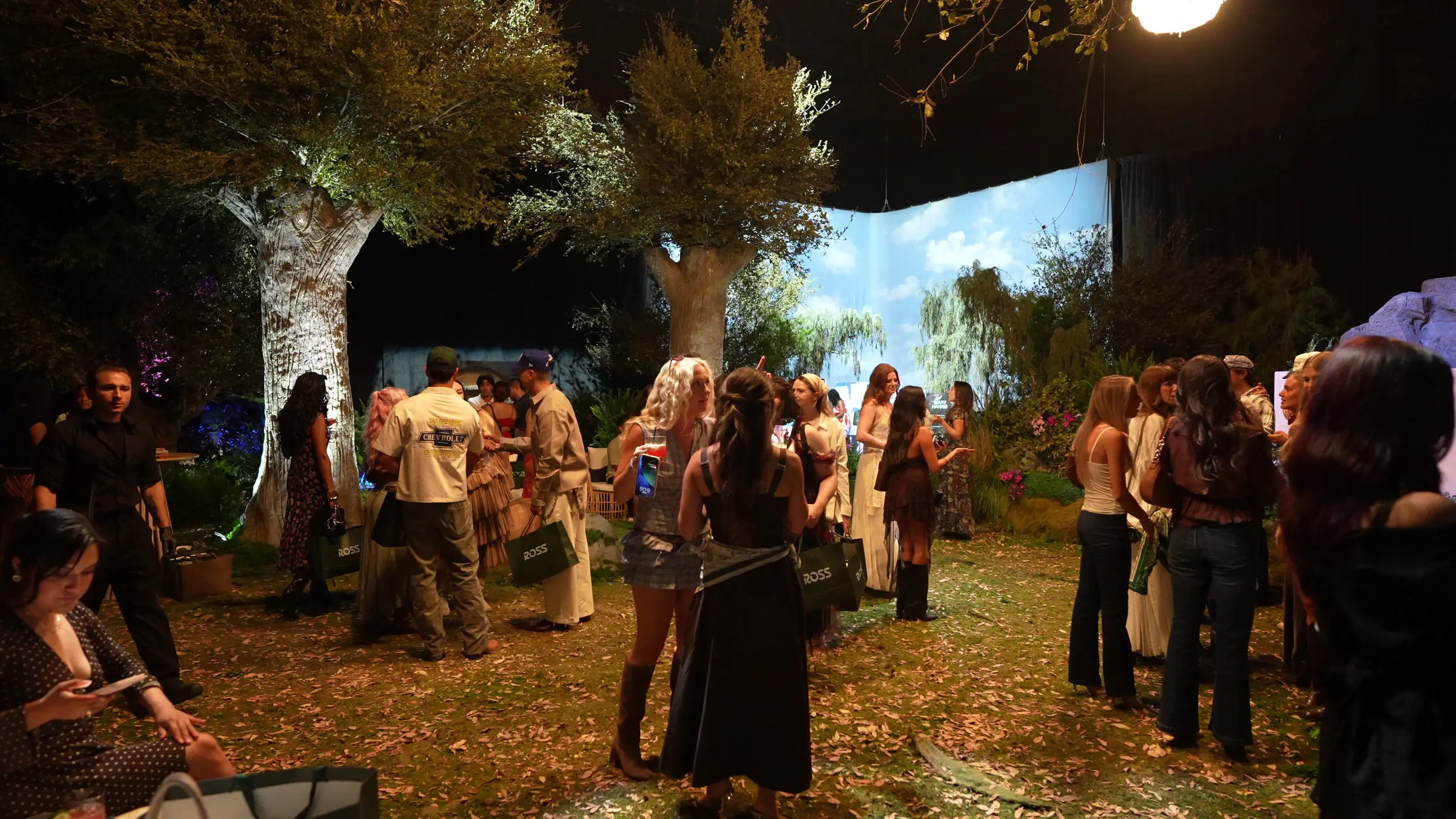 Guests carrying green Ross bags across the leaf covered turf, the two sculpted trees lit from below and the projected sky wall glowing behind them.