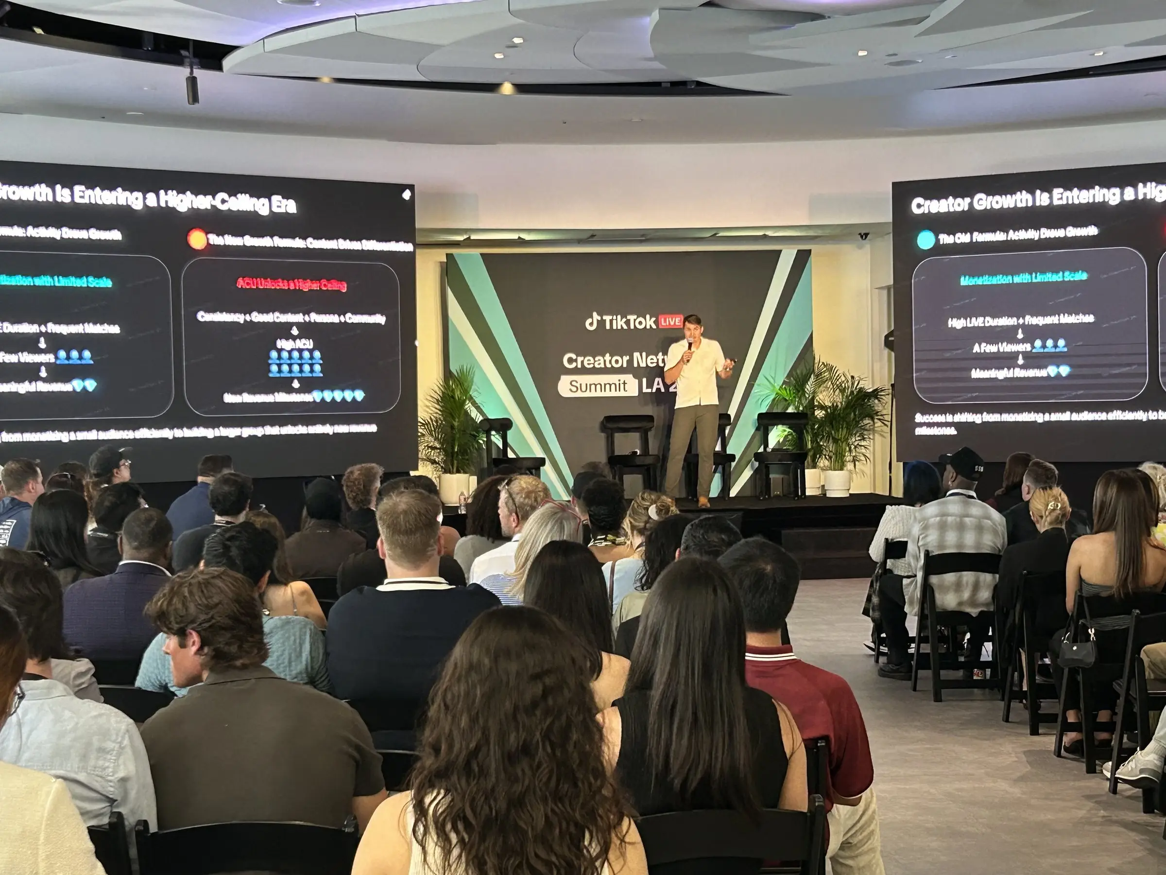 A speaker on the small stage between two content screens, seen from the back of the seated room.
