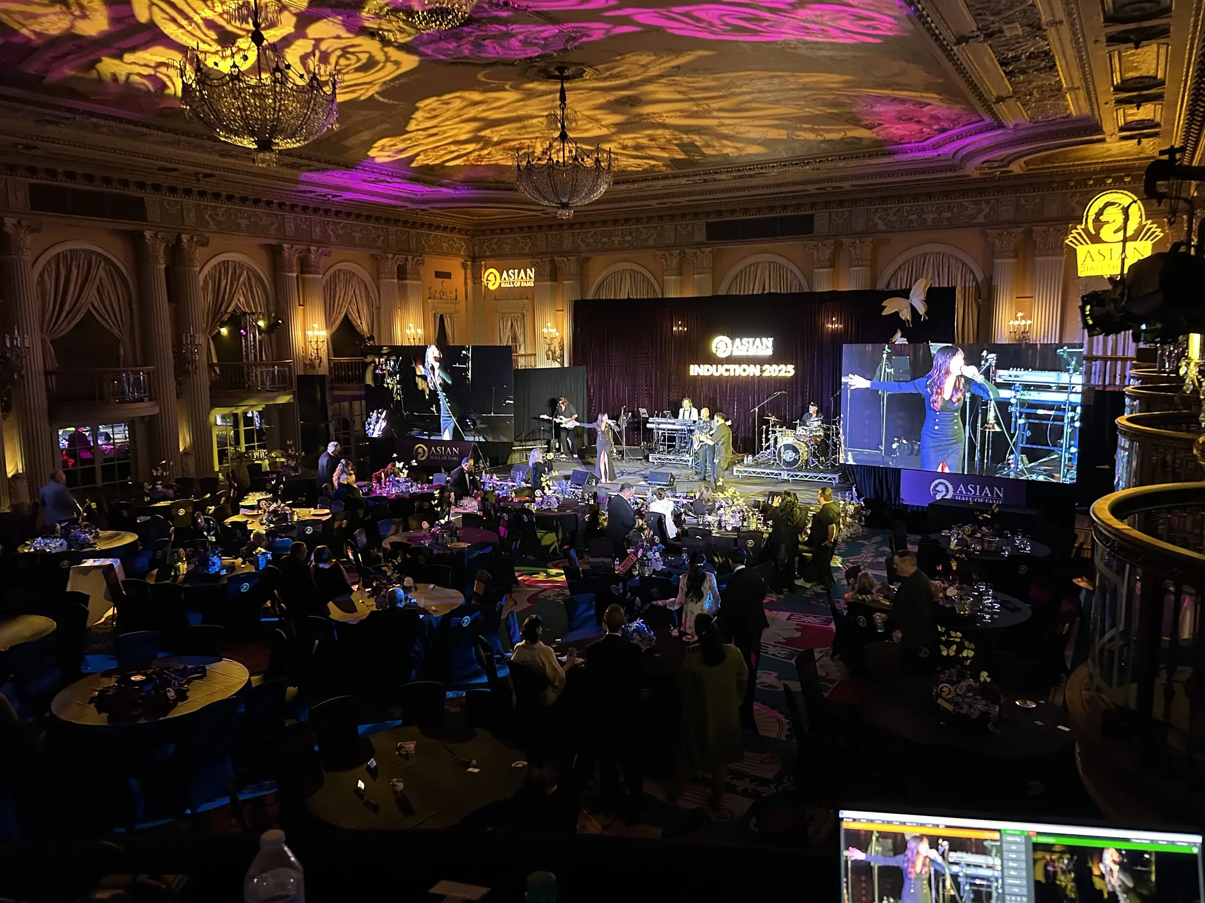 Warm gold light on the ceiling plaster, guests moving between the tables, and a band playing under stage wash.