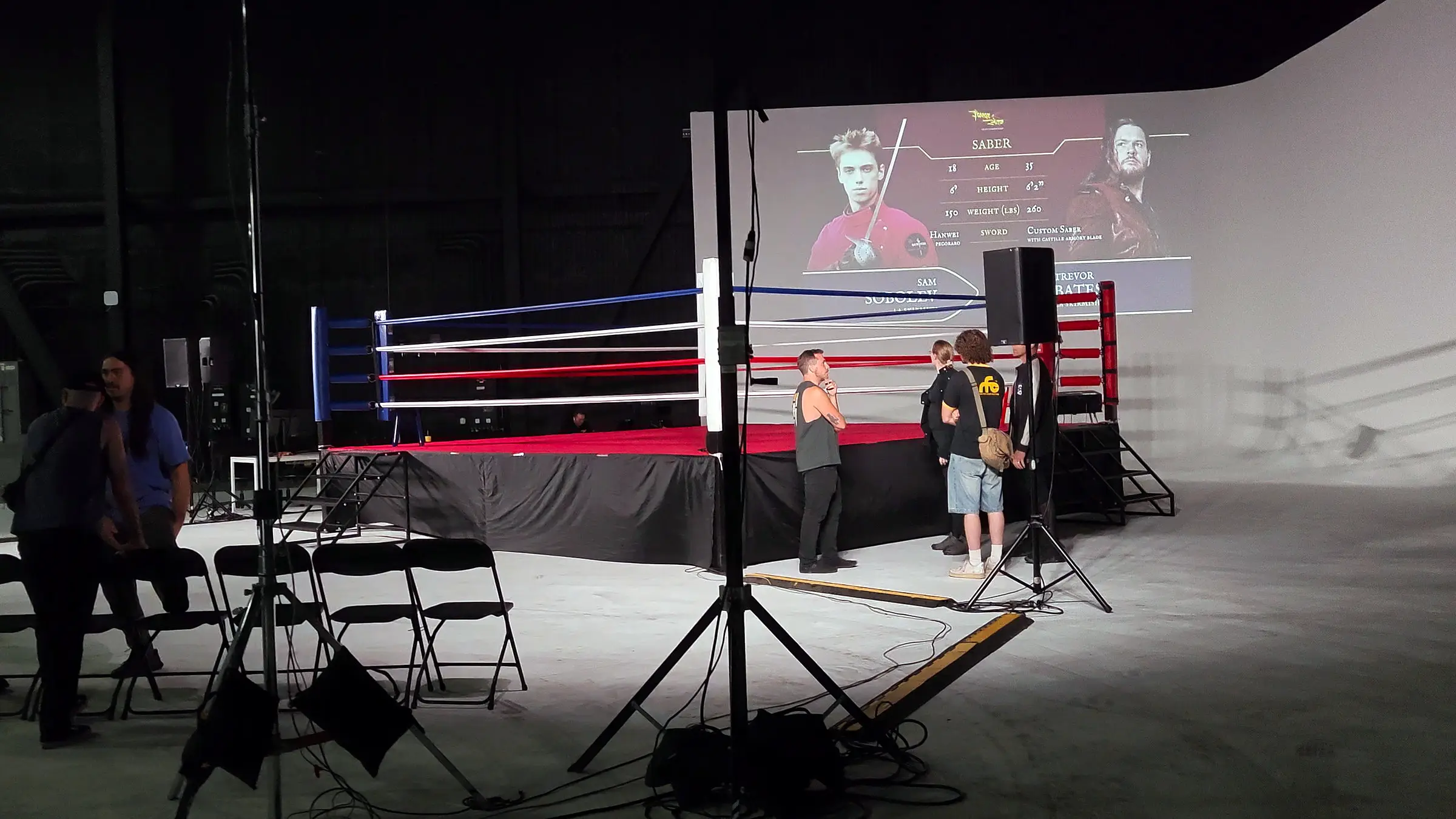 The ring before doors with a saber bout card projected on the cyclorama, light stands and cable runs across the floor, and crew standing at the apron.