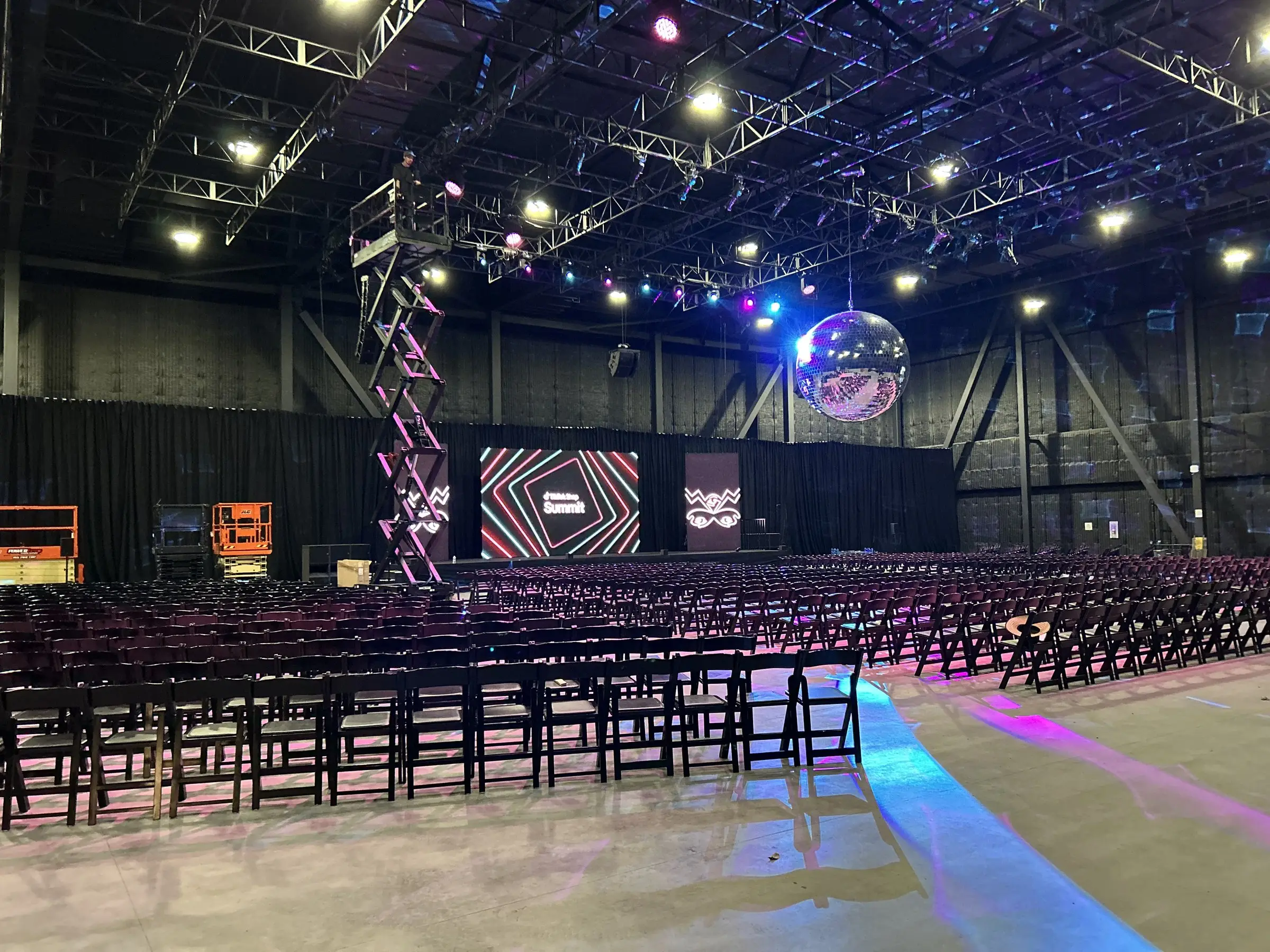 Load-in in the same hall, a lift parked under the truss, LED panels stacked at the stage, and chairs set in rows on bare floor.