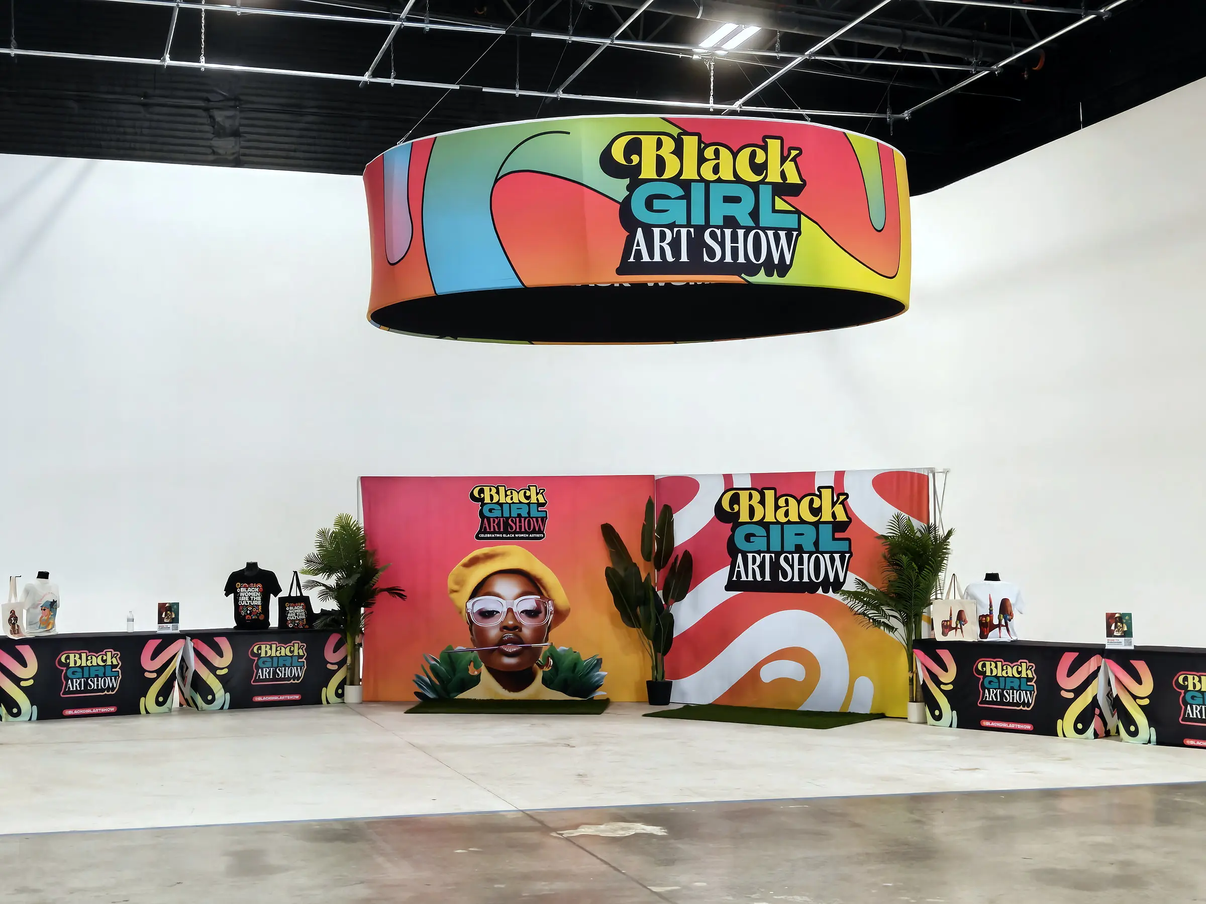 The merch counters and the printed photo wall set against the white cyc, with a round Black Girl Art Show banner hung above them.