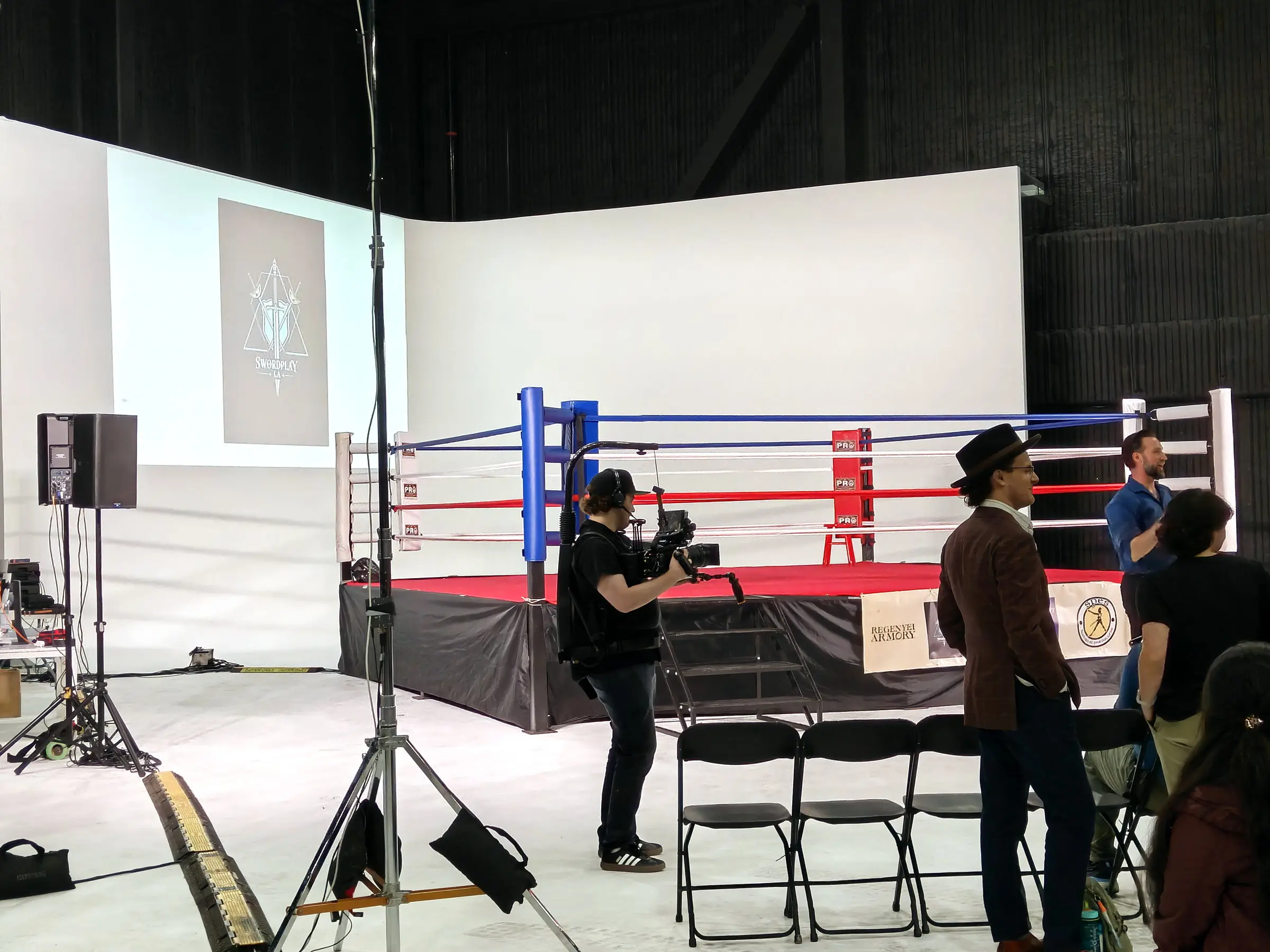 A camera operator on a gimbal shooting the empty ring while the ring announcer in a top hat waits at the ropes.