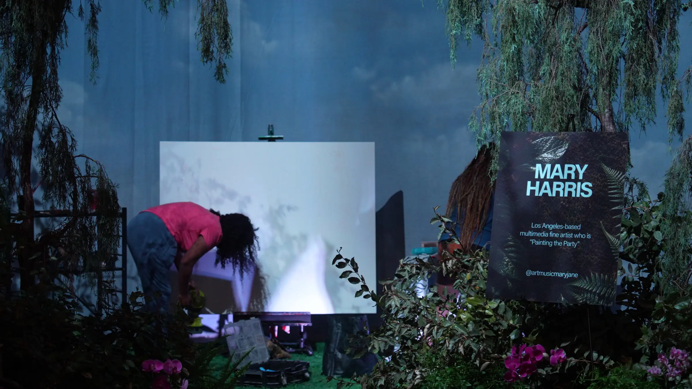 The live painting station, an artist bent over a blank canvas on an easel under the willow branches, next to a lit sign for the artist Mary Harris.