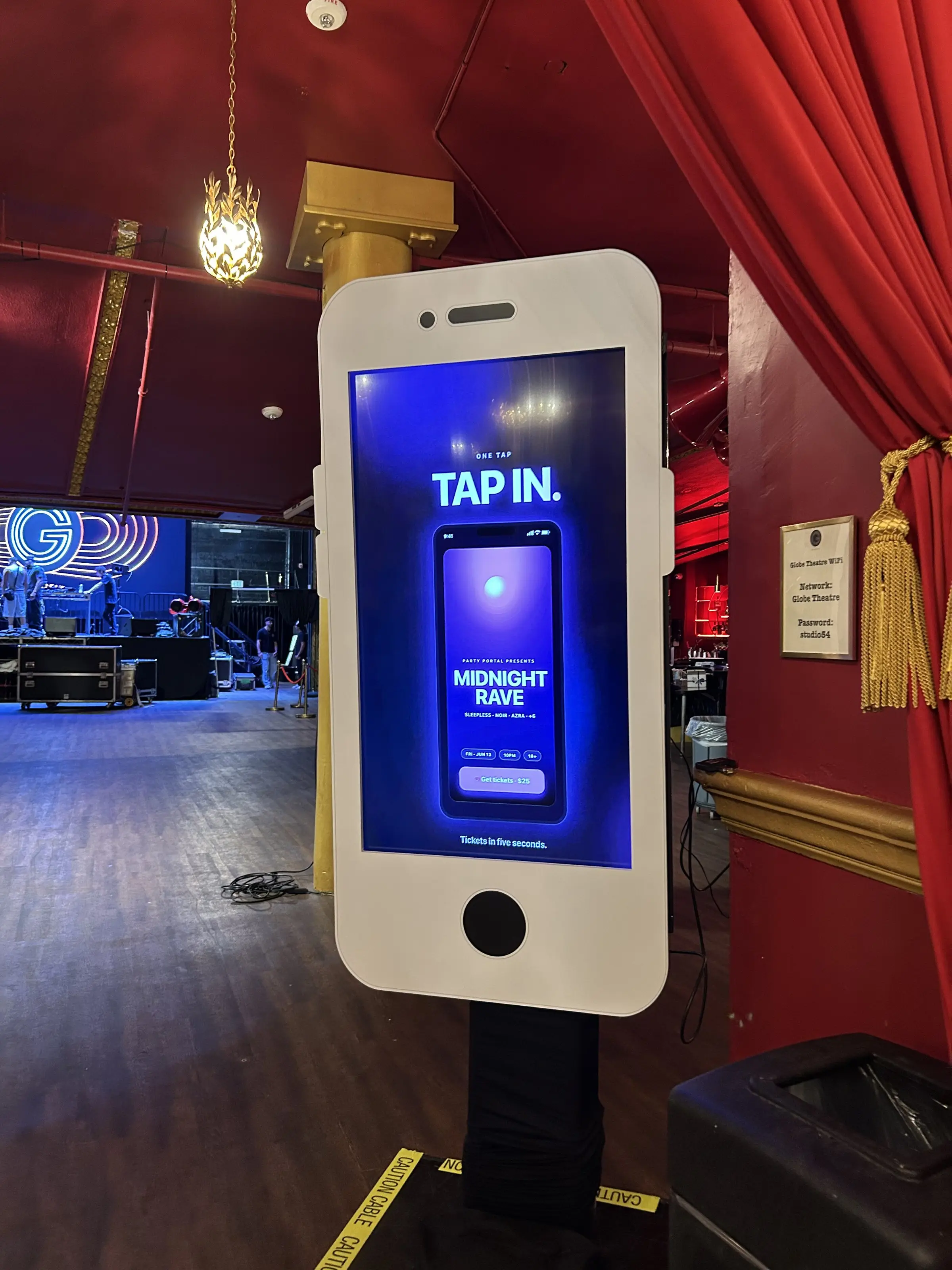 An oversized phone shaped kiosk in the lobby, its screen reading Tap In, with red drapes behind it.