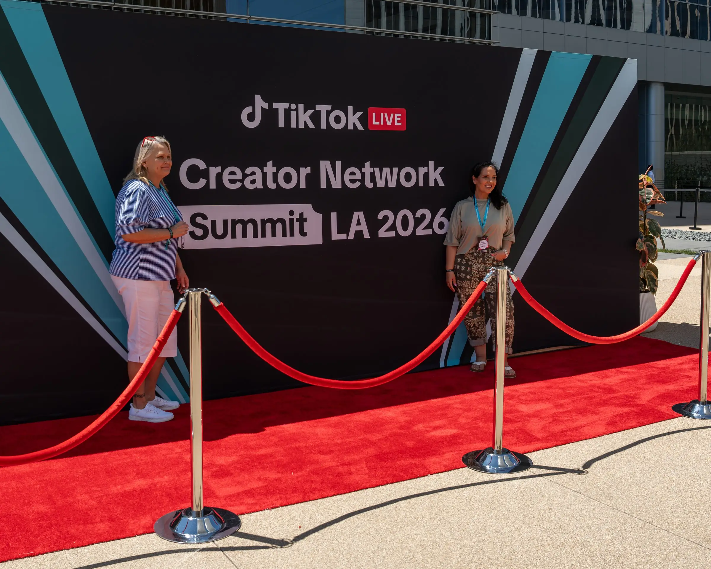 The red carpet outside in daylight, two women standing at the step and repeat behind a red rope line on chrome stanchions.