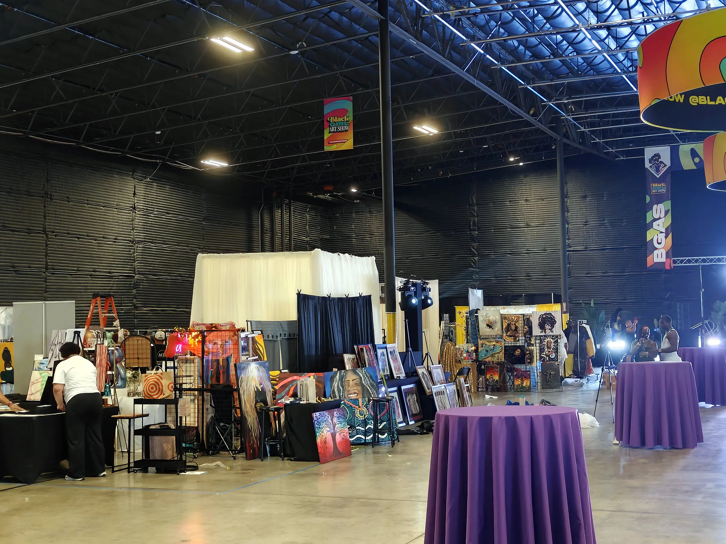 Artists dressing their booths, canvases propped on stands and grid walls, with purple cocktail tables set across the floor beside them.