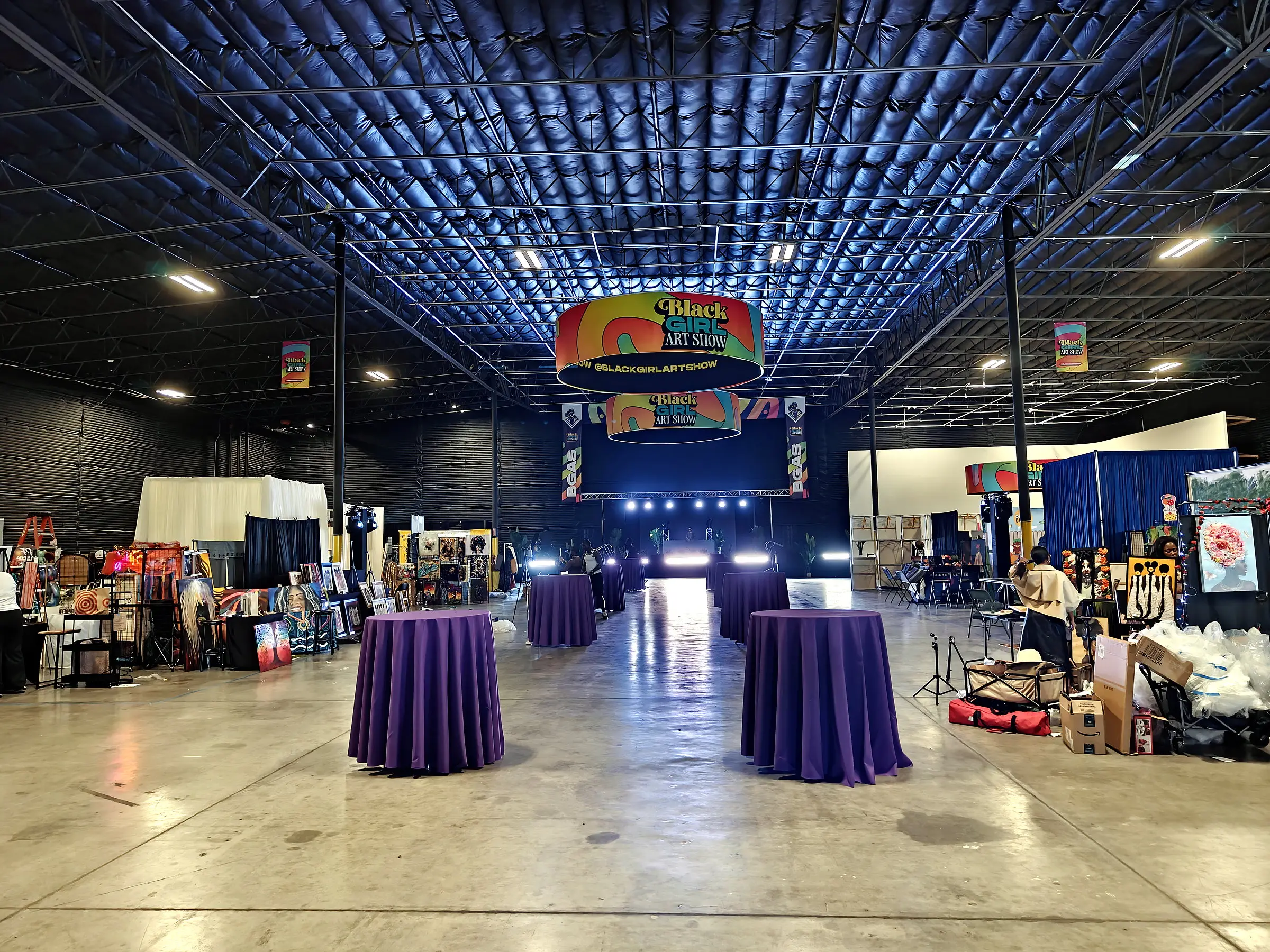 The hall under house light, art booths down both sides, two round Black Girl Art Show banners hung over the floor, and the stage lit at the far end.