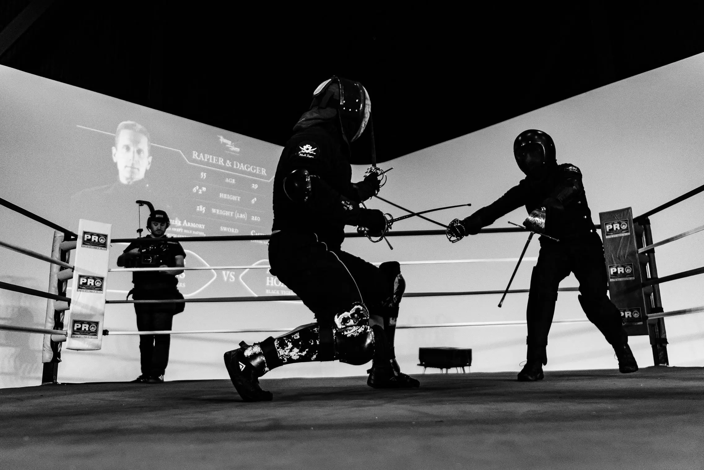 Two masked fighters lunging at each other in the ring, the bout card projected on the wall behind them and a camera operator shooting from the corner.