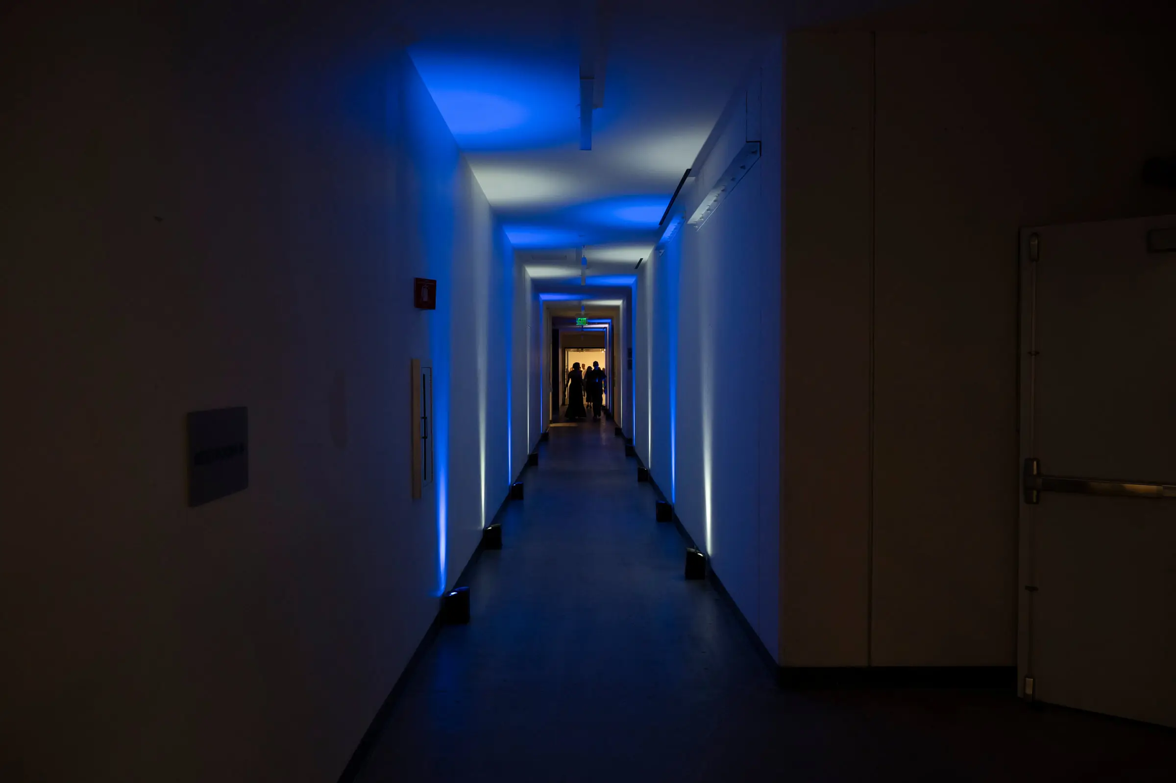 A corridor washed blue from uplights on the floor, with a lit doorway and two small figures at the far end.