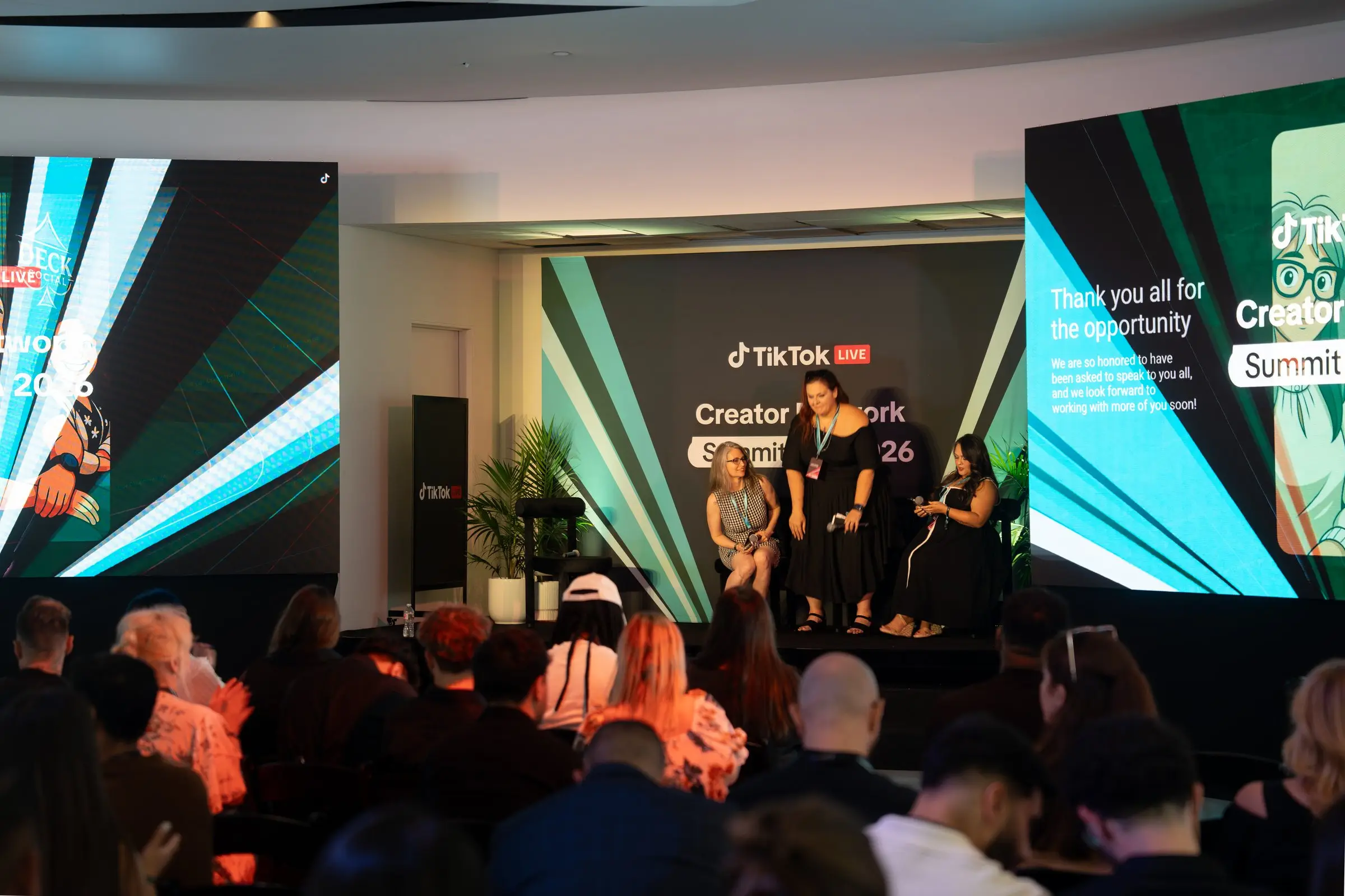 Three speakers standing on stage with microphones while both LED walls run graphics, seen over the heads of the seated audience.