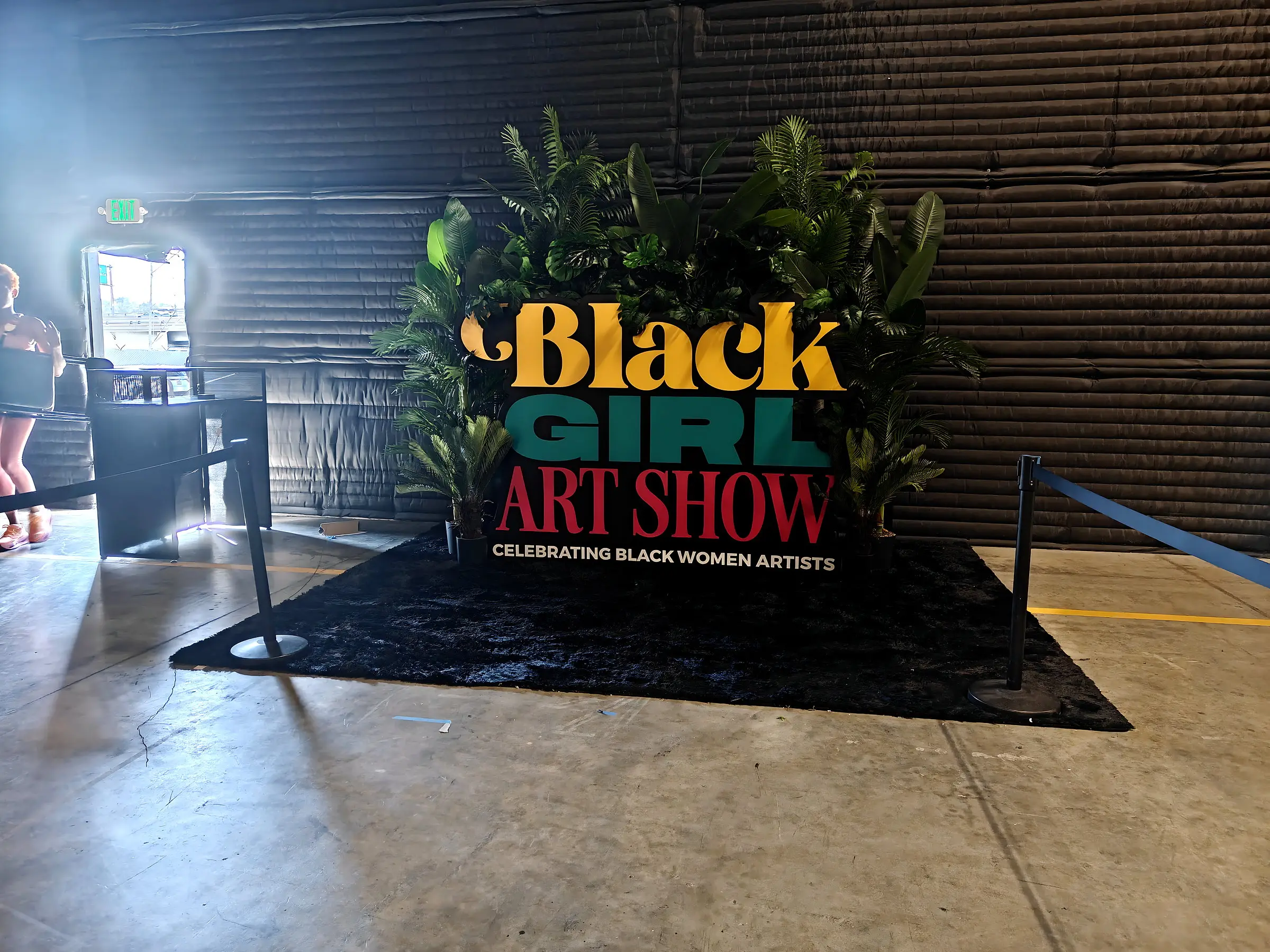 The Black Girl Art Show entrance sign on black turf with plants behind it, rope stanchions in front, and daylight coming through the door.