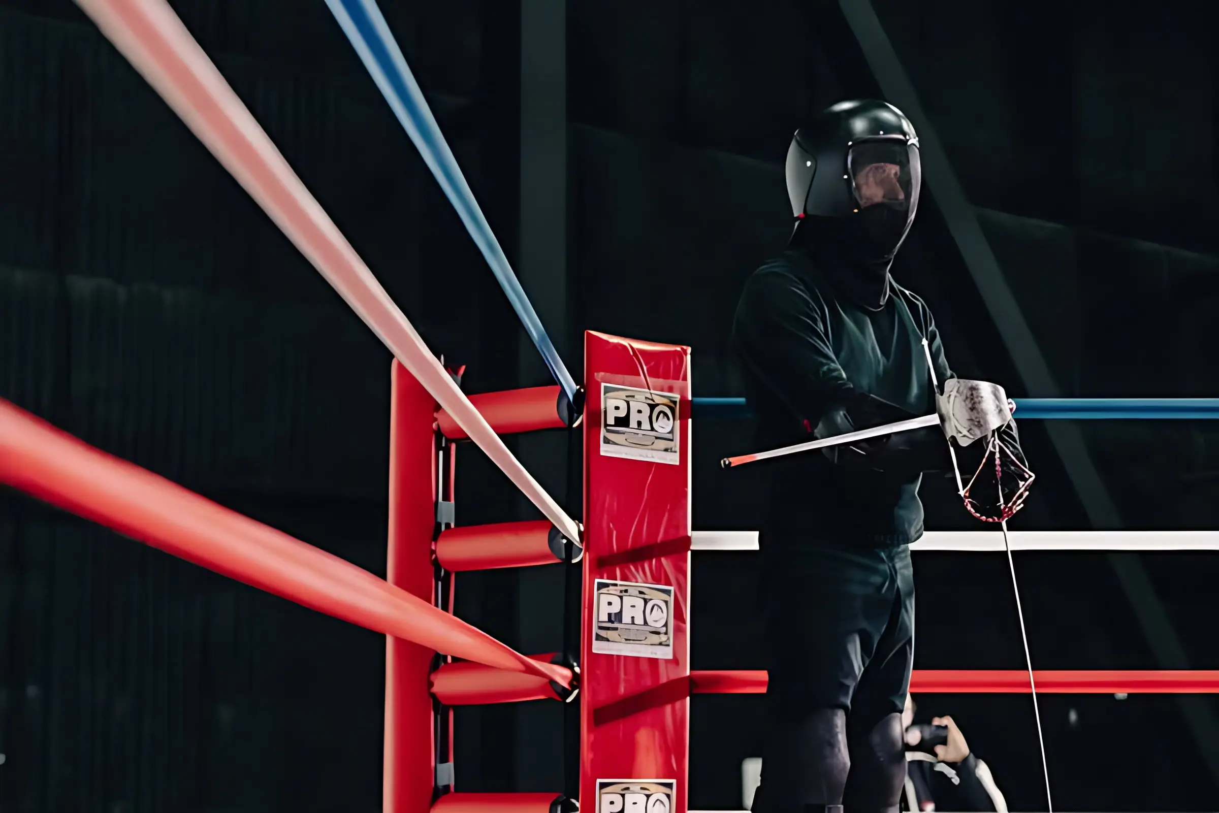 A fighter in a fencing mask and gauntlets standing at the red ring post with a sword in hand.
