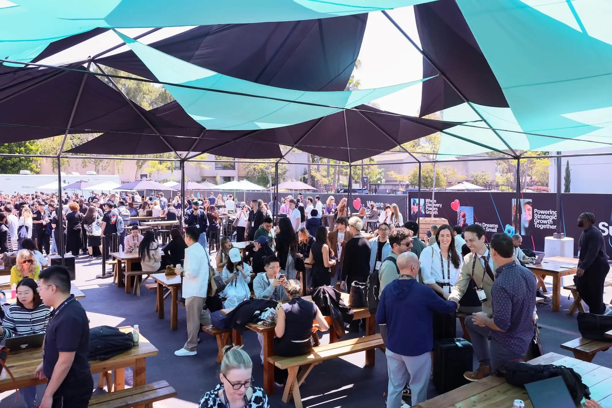 Black and teal shade sails over an outdoor courtyard, picnic tables underneath, and a crowd filling the space.