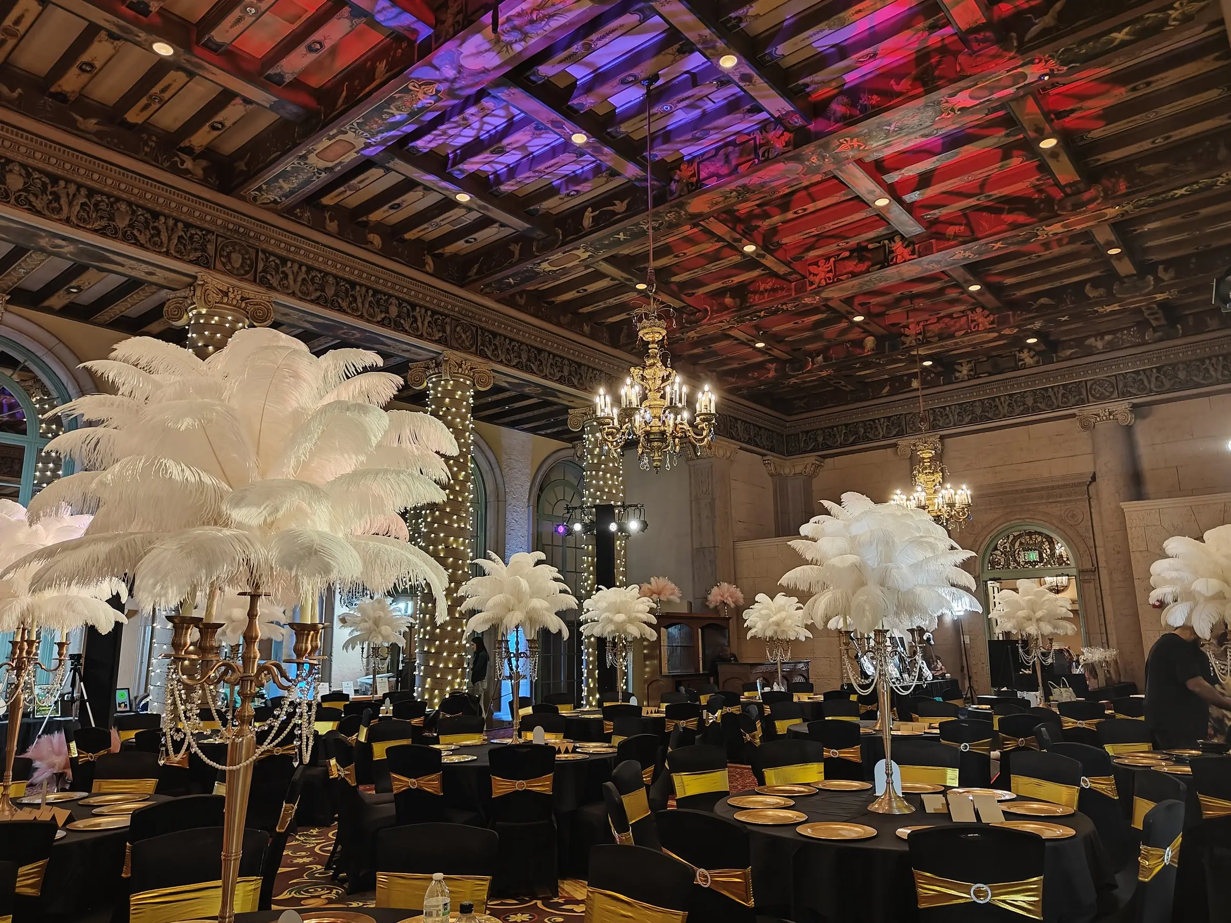 The painted ballroom ceiling washed red and purple above the string lit columns, with white feather centerpieces and gold set tables filling the floor below.