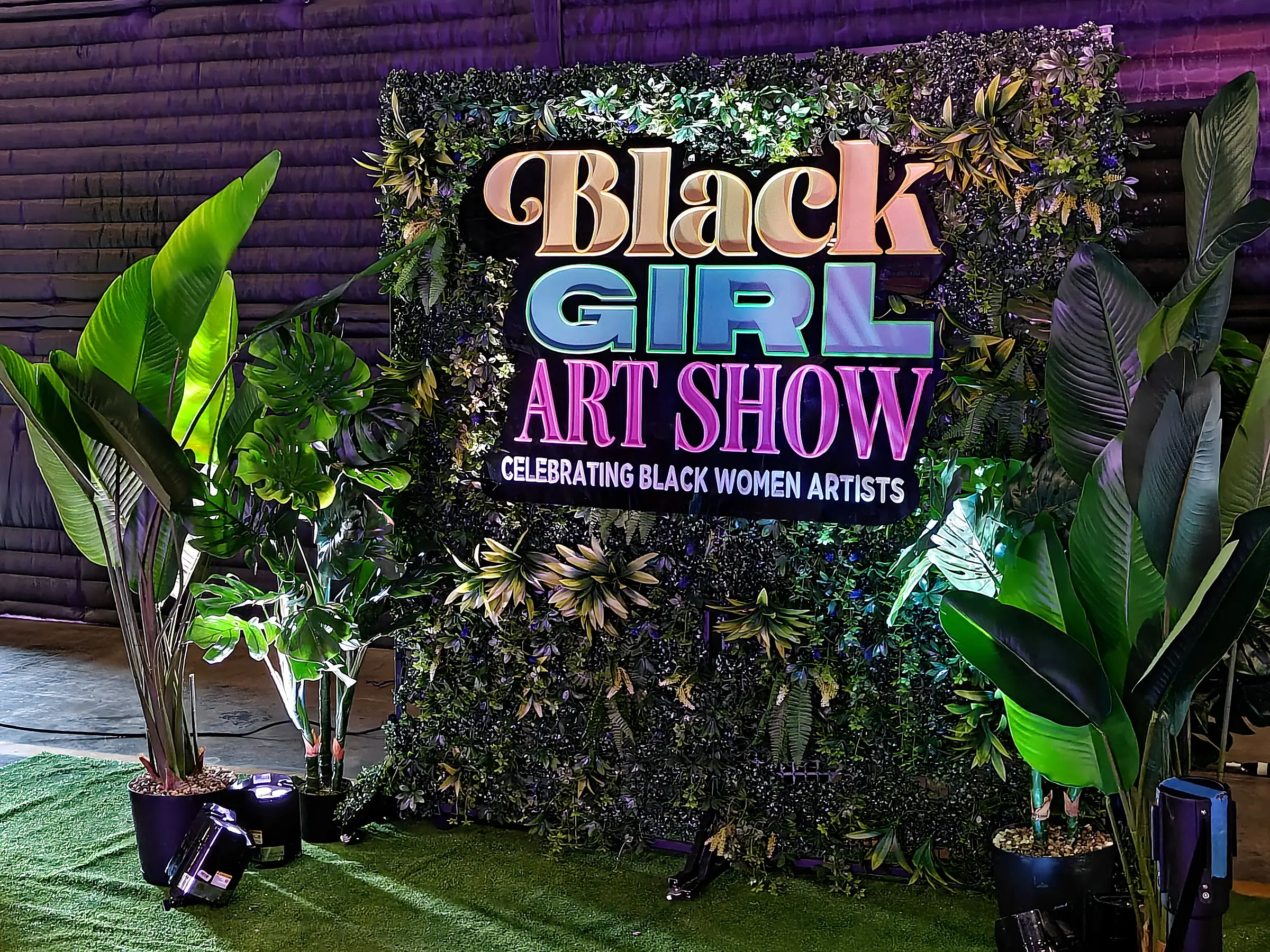The greenery step and repeat lit from the floor at night, the show sign mounted in the middle of it and potted plants on the turf at both sides.