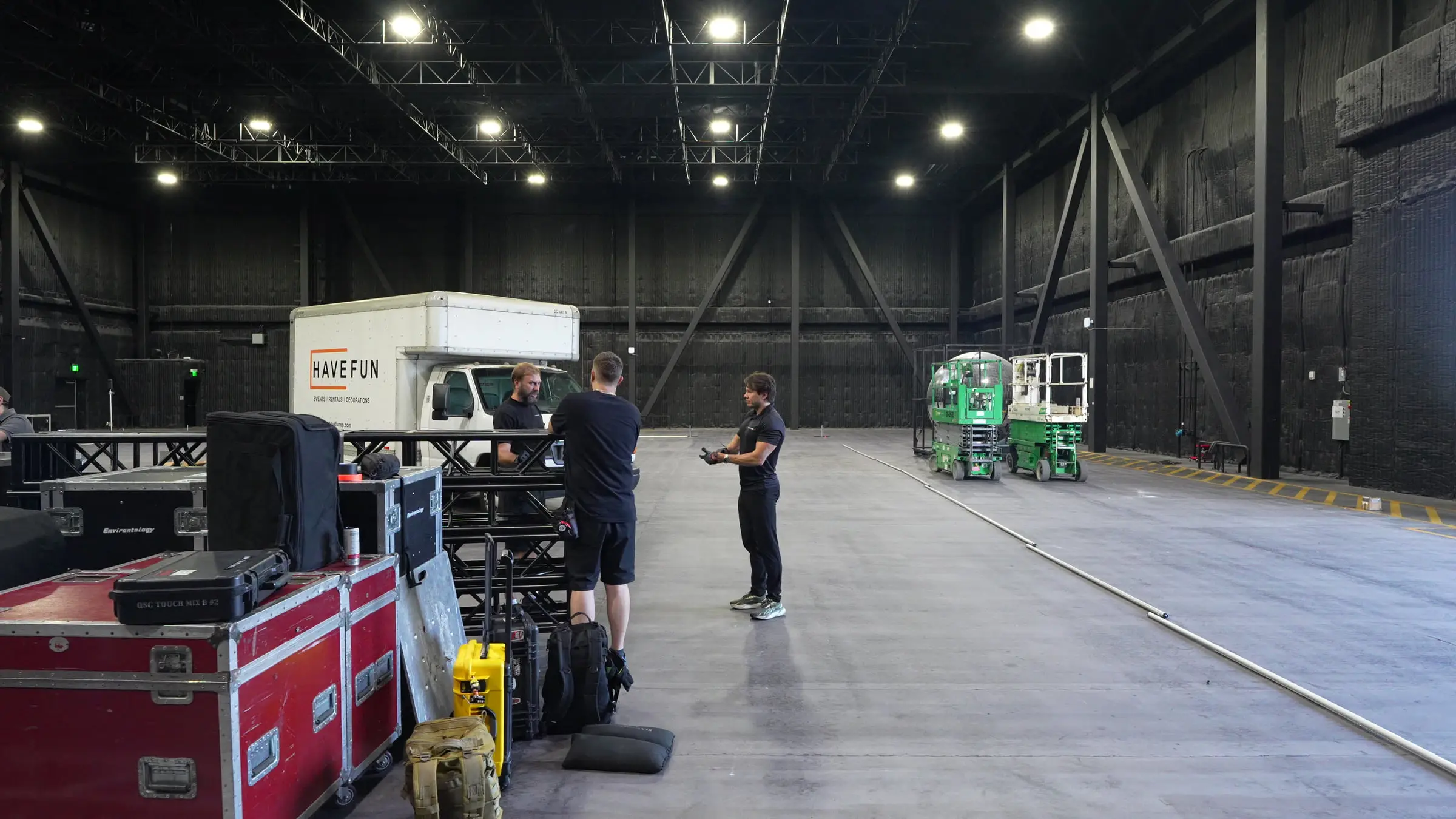 The empty soundstage during load-in, a box truck parked on the concrete floor beside stacked road cases, truss sections, and two scissor lifts.
