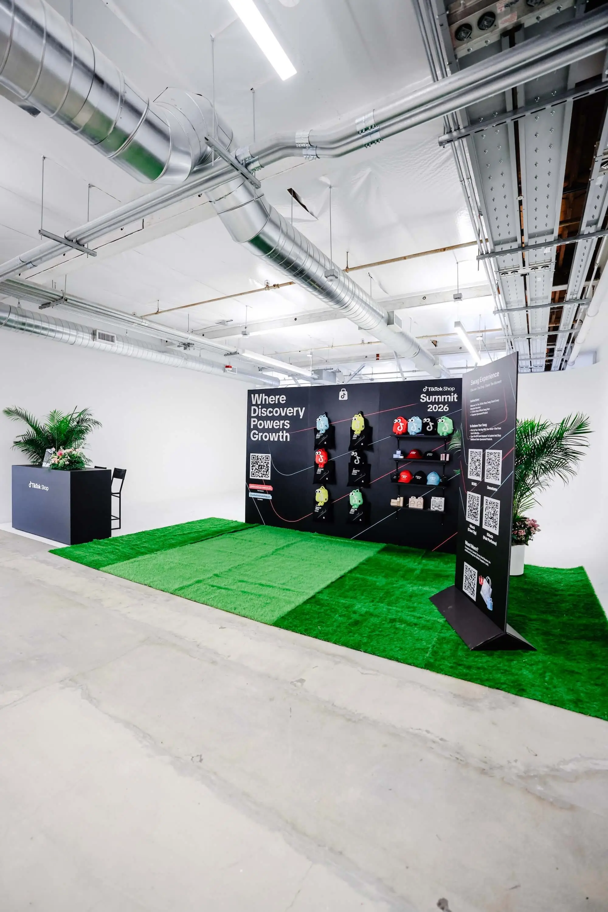 A booth built on a square of green turf, a dark backdrop reading Where Discovery Powers Growth, and a counter beside potted palms.
