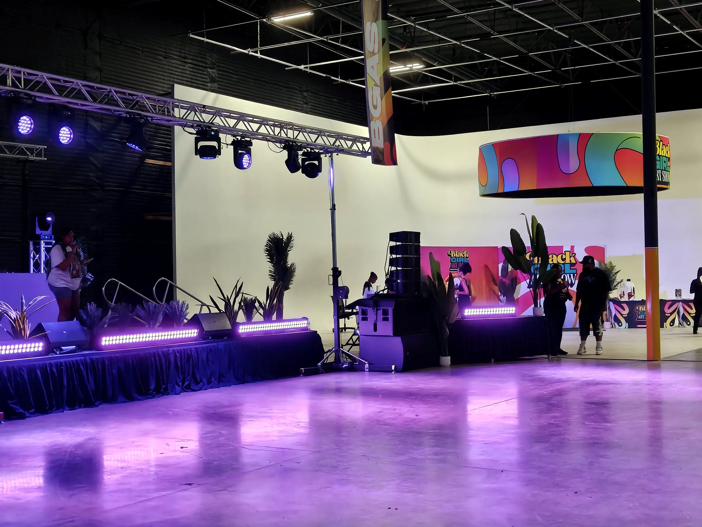 The stage from the floor, LED bars along the deck, a speaker stack beside it, and moving lights on the truss washing the room magenta.