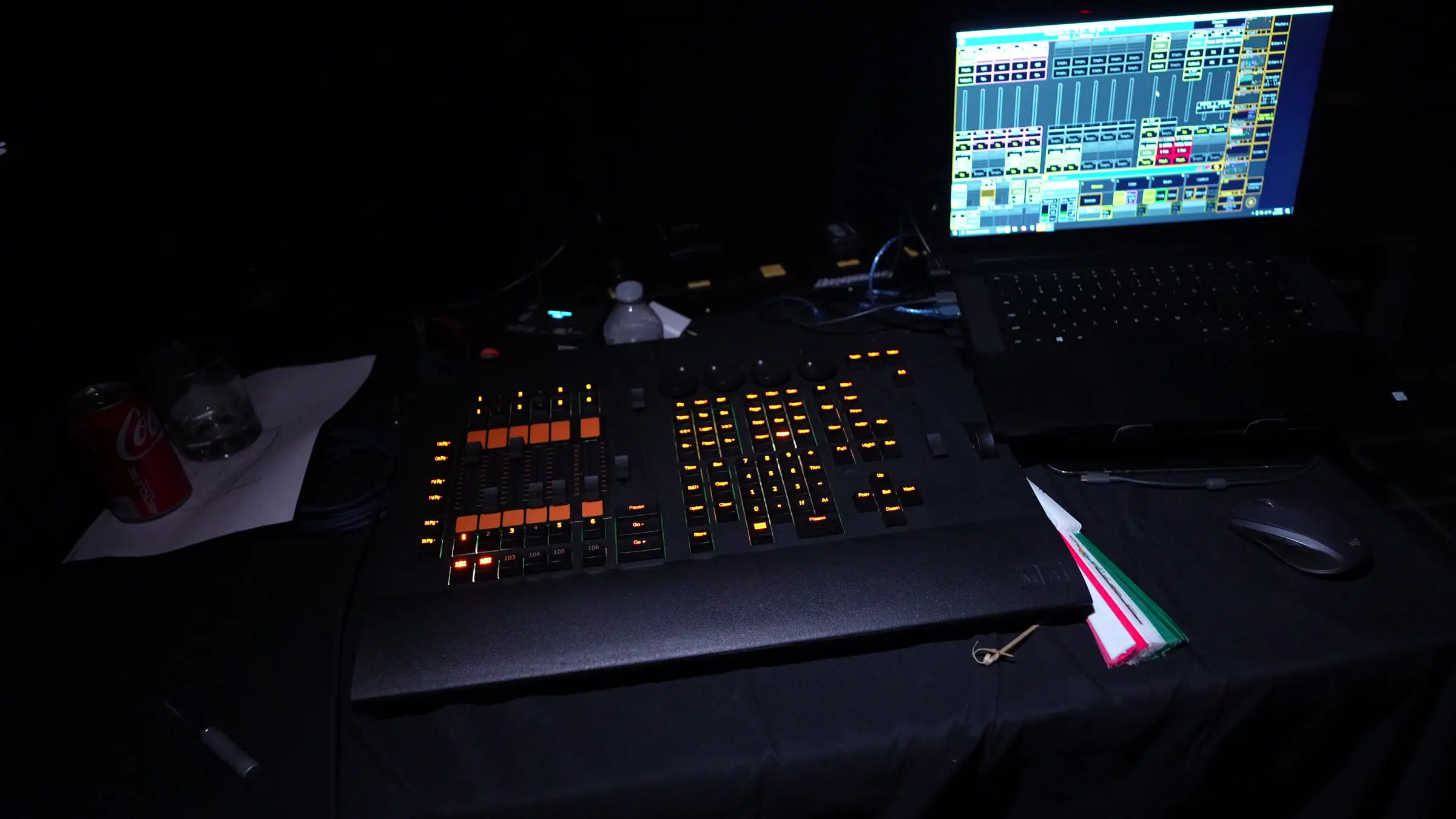 The lighting console lit by its own keys on a draped table in the dark, with a laptop running the show file beside it.