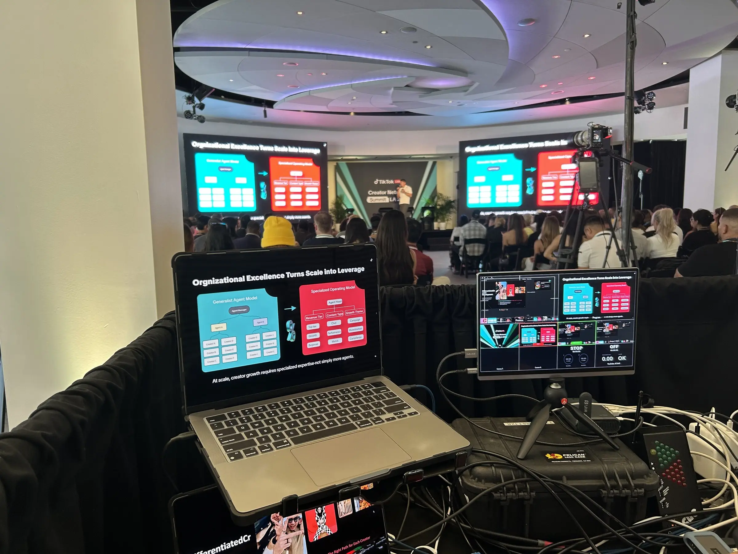A laptop running the presentation slide beside a multiview monitor on the tech table, with the seated audience and both LED walls carrying the same slide beyond.