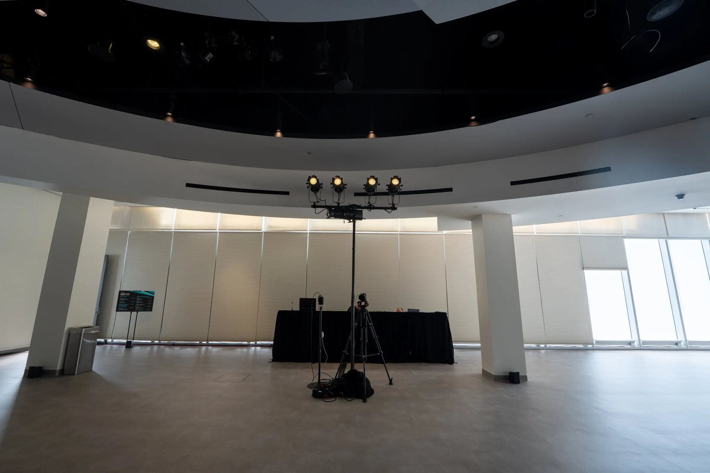 A light tree with four fixtures standing in the empty room, a draped table behind it and the curved wall of closed blinds under the black ceiling.