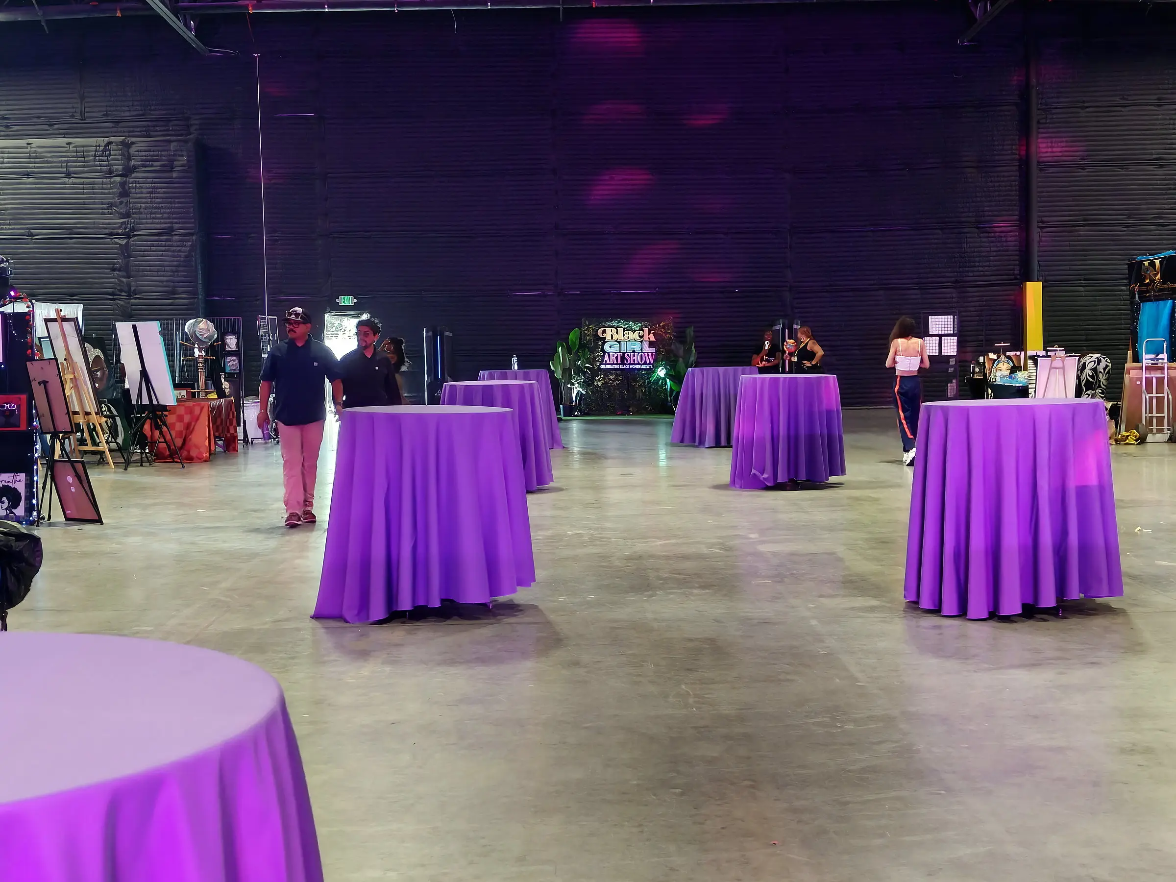 Guests walking between purple cocktail tables under magenta wash, art easels along the left wall and the greenery sign lit at the back of the room.