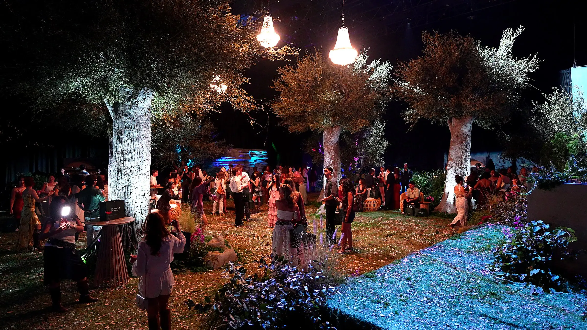 The forest from the far side of the room, three sculpted trees lit from below, two chandeliers hanging over the crowd, and the stacked stone wall washed blue behind them.