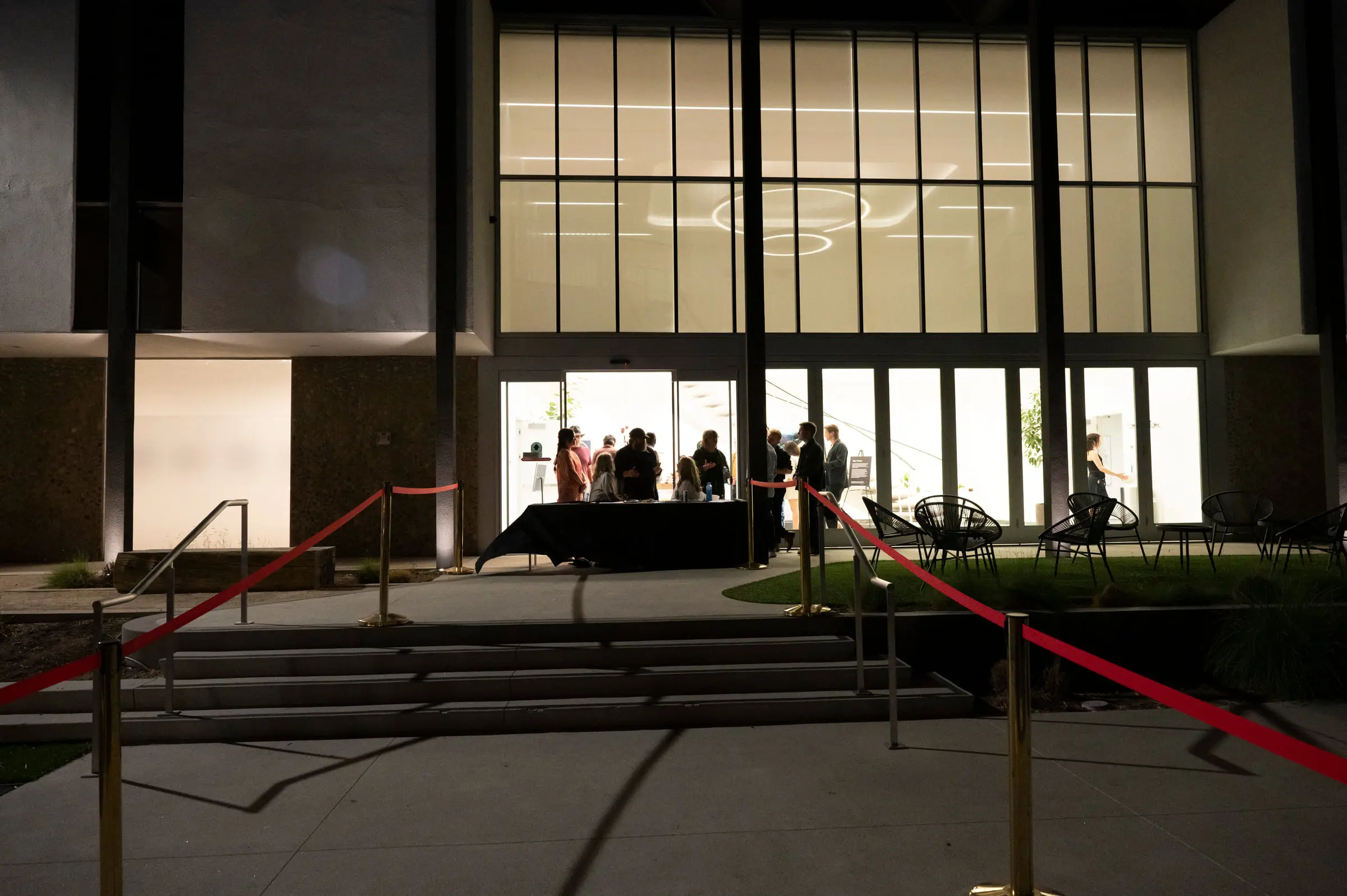 The check in table at the lit entrance after dark, guests in silhouette in the doorway and a red rope line across the steps.