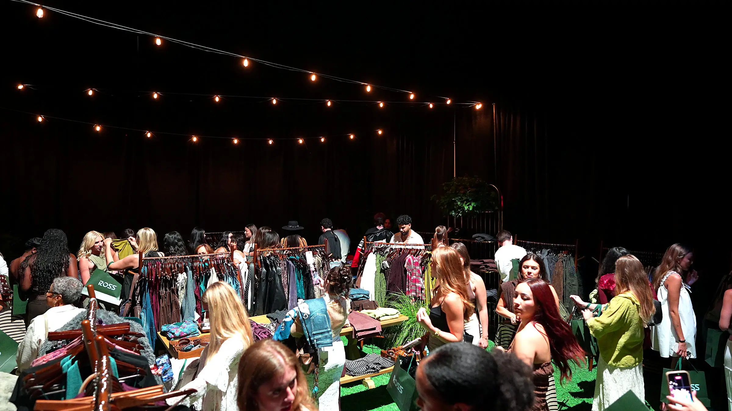 Guests working through the clothing racks and the folded stock on the tables in the gifting suite, with strings of bulbs hung across the black drape above them.