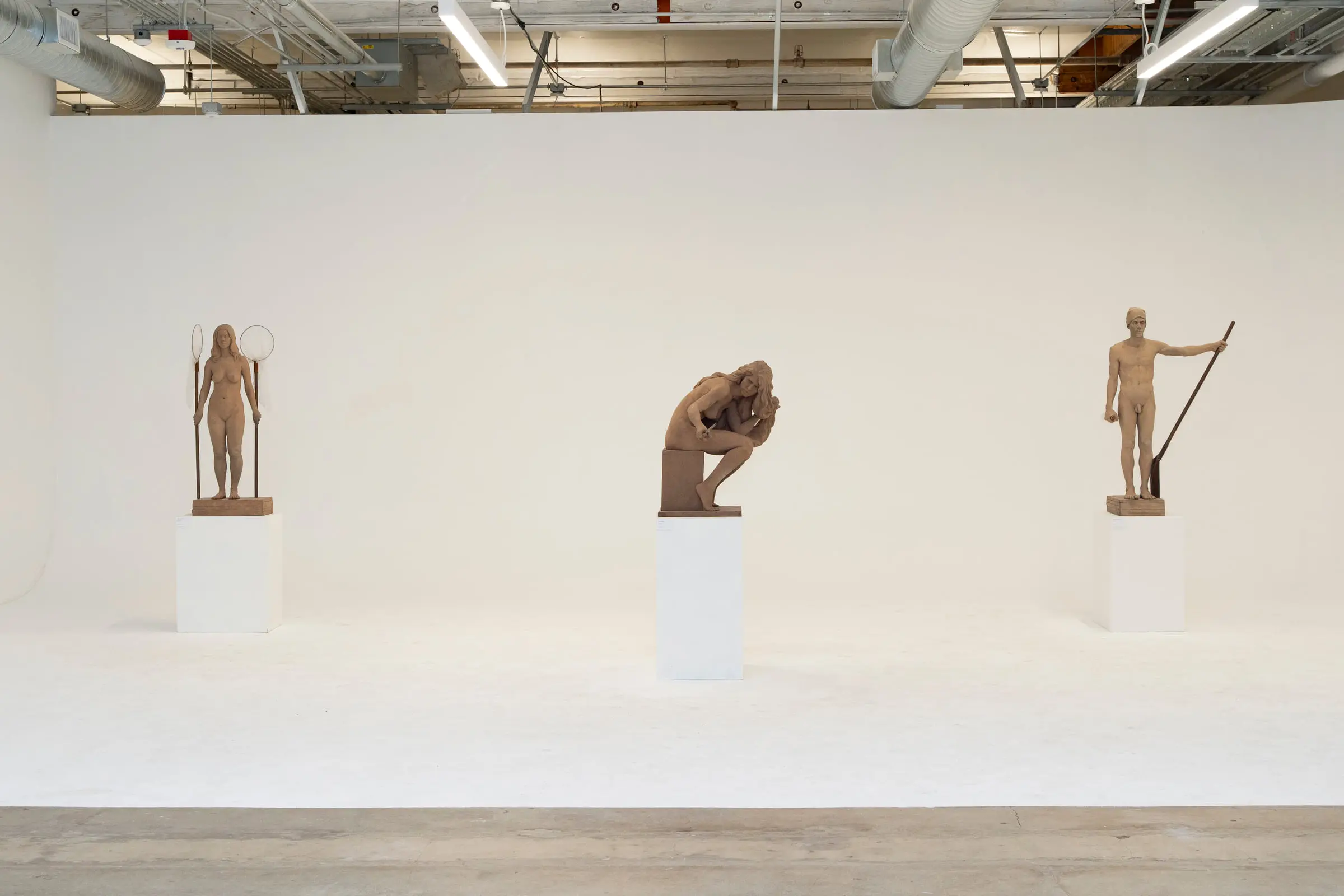The white cyclorama room with nobody in it, three carved figures on plinths spaced along the curved wall under the exposed ducts and strip lights.