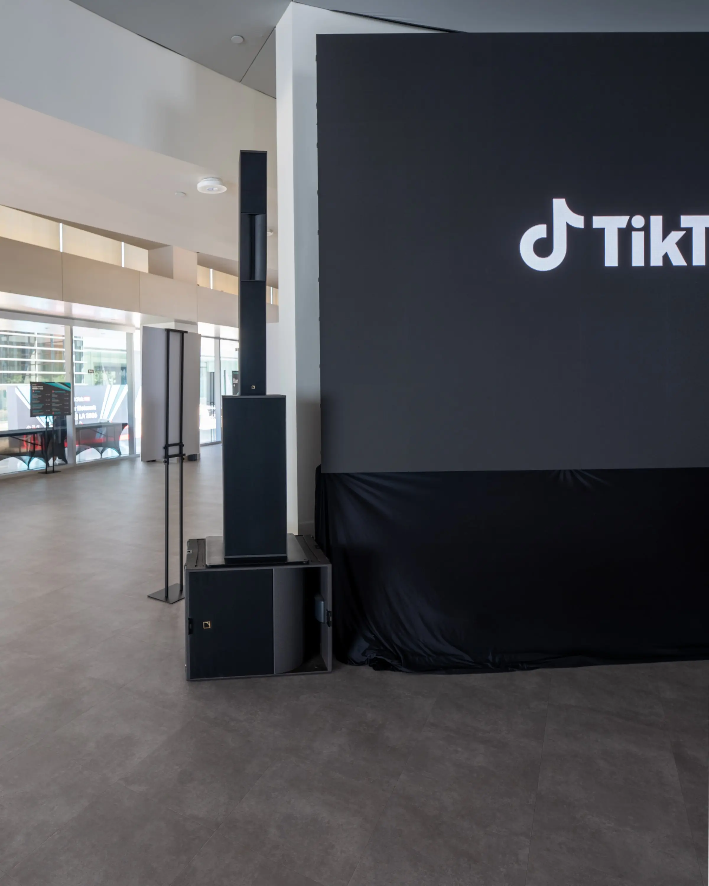 A speaker column stacked on a subwoofer beside the LED wall showing the TikTok Live logo, with the bare floor and the glass lobby behind it.