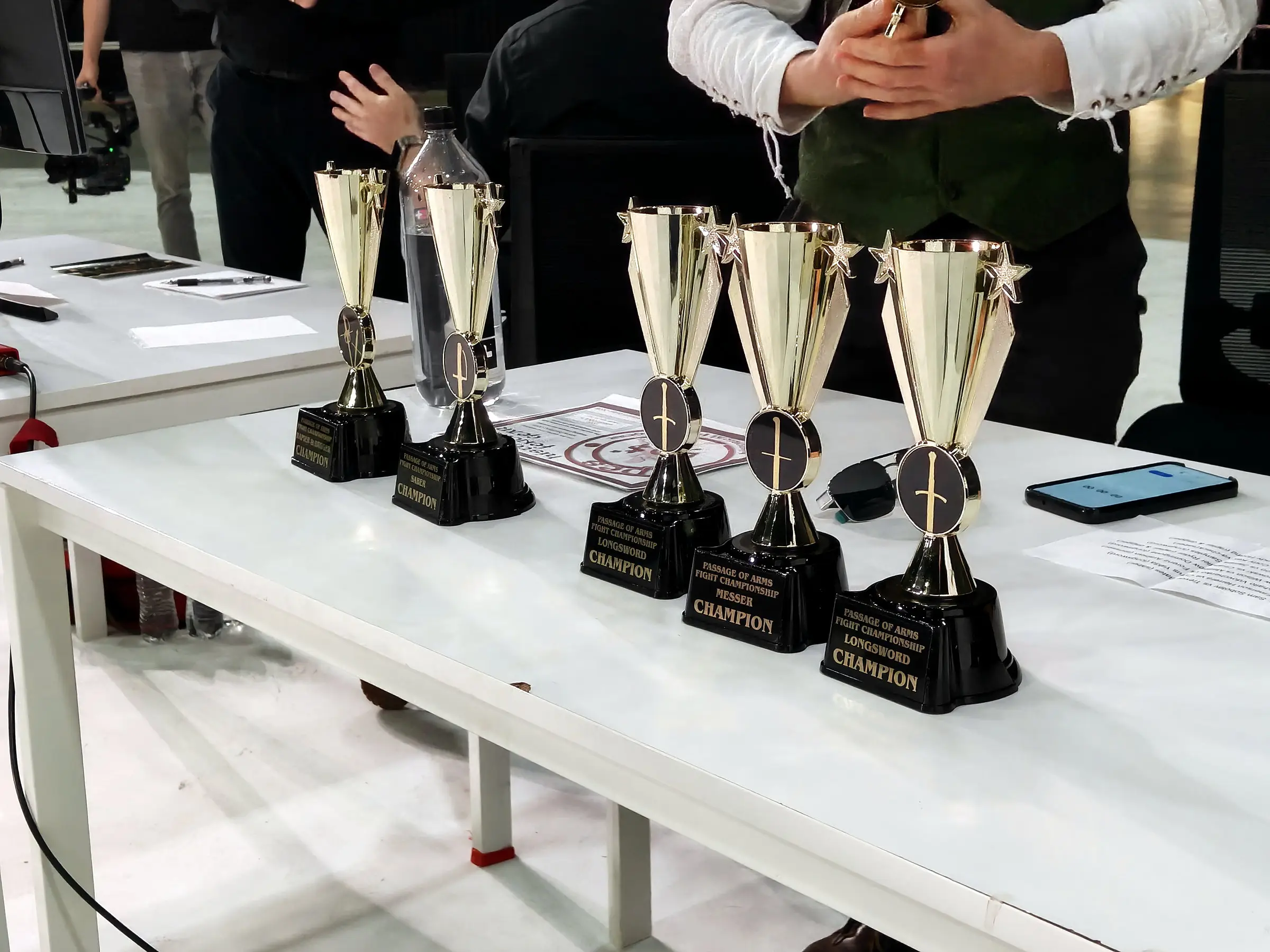 Five trophies on a white table, their plates reading Passage of Arms Fight Championship with the saber, longsword and messer champion titles.