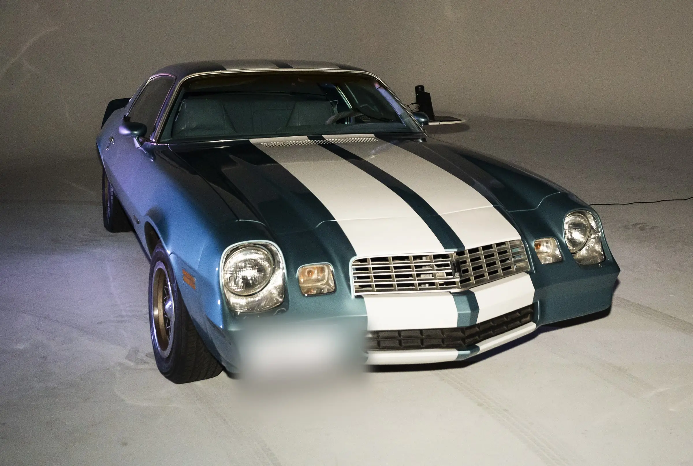 A blue coupe with two white stripes over the hood parked on the bare studio floor, lit from the front with the wall dark behind it.