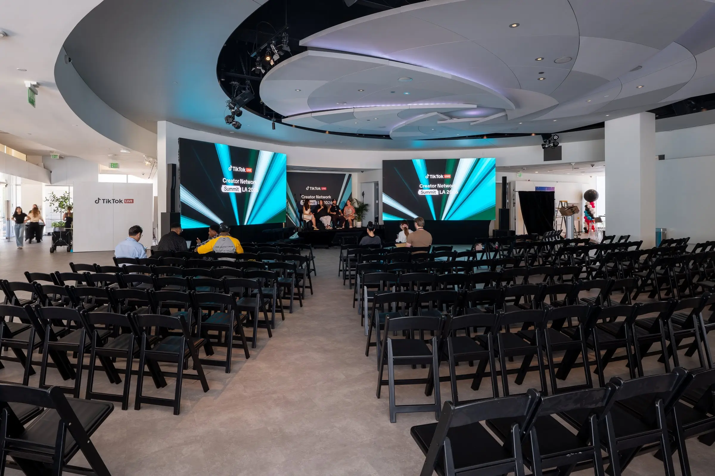 Rows of black folding chairs with a center aisle facing the stage between the two LED walls, a few guests seated while the panel talks under the blue lit ceiling.