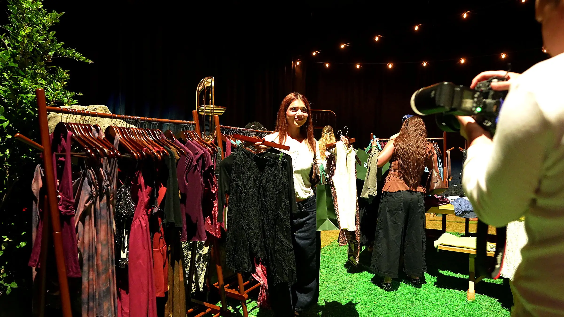 A guest holding a jacket up off the rack in the gifting suite while the videographer films her from the right, string lights running across the drape behind.