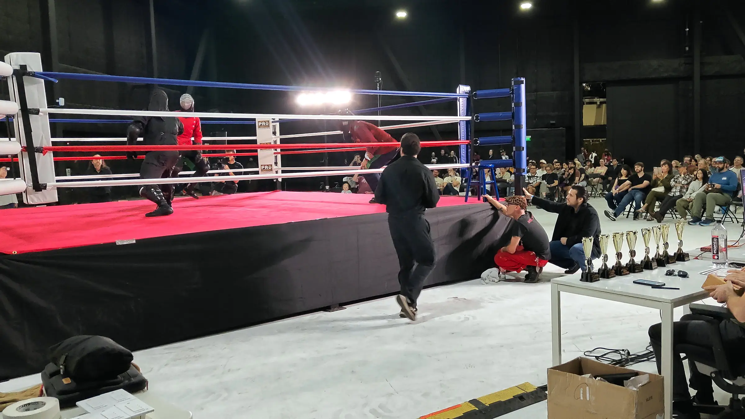 The room working, a bout in the ring, the crowd seated along the far wall, and the trophy table and production desk in the foreground.